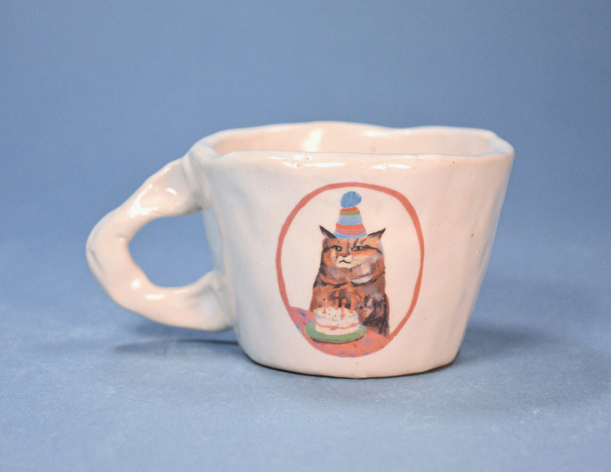 White ceramic mug with a cat wearing a party hat and birthday cake design on a blue background