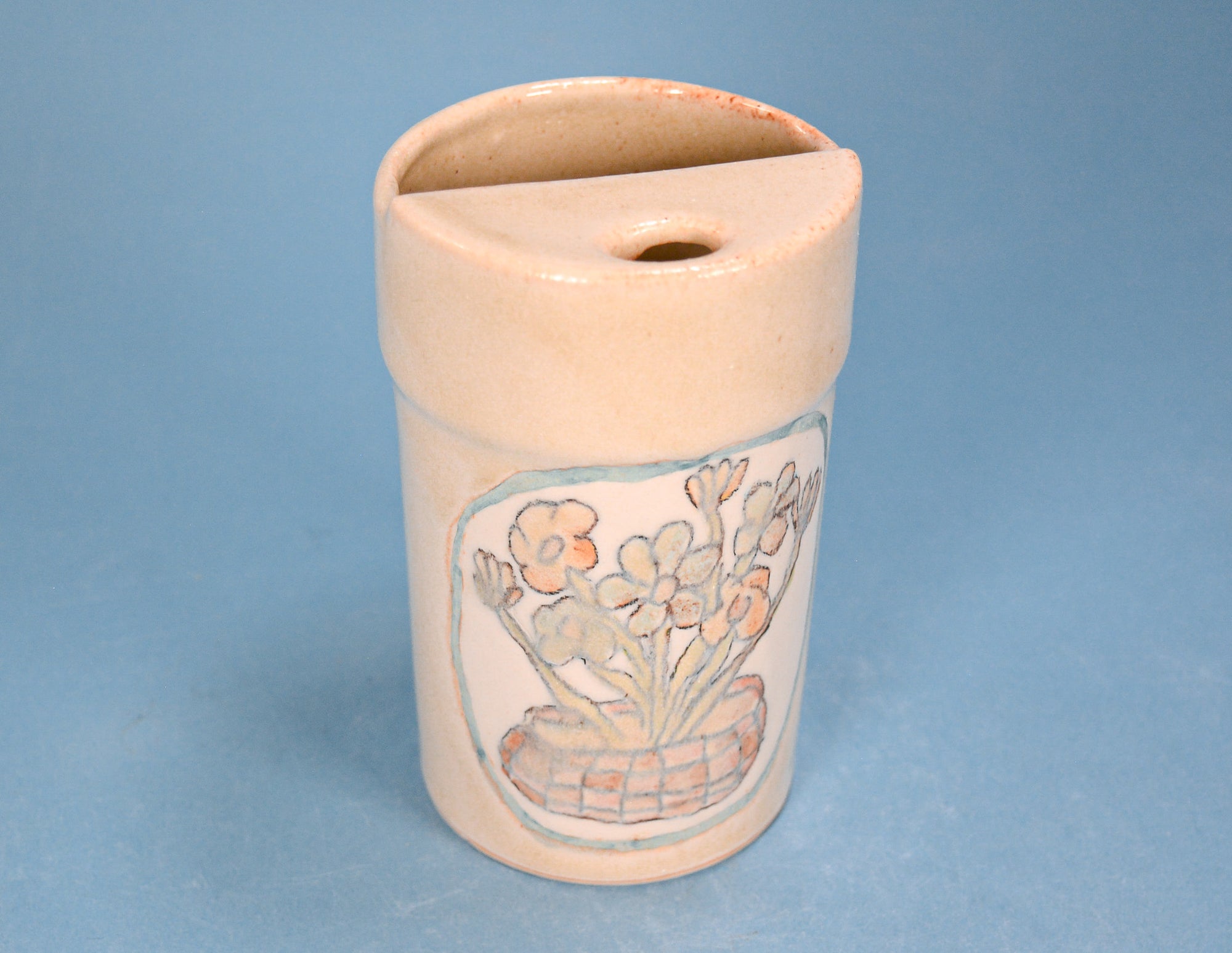 A small ceramic container with a slanted lid and a painted floral design on the front, photographed against a solid blue background.
