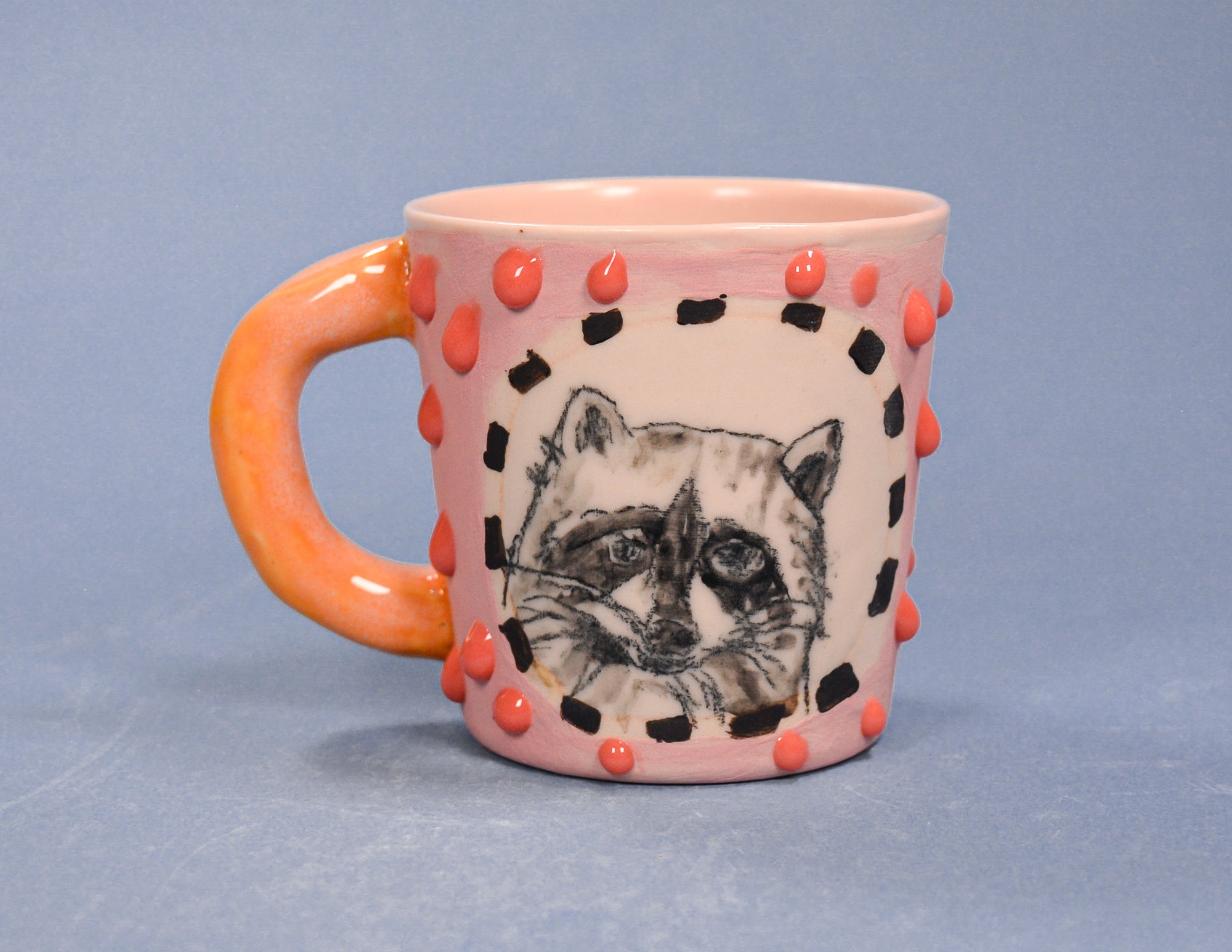 Pink mug with orange handle and raccoon illustration on a light blue background