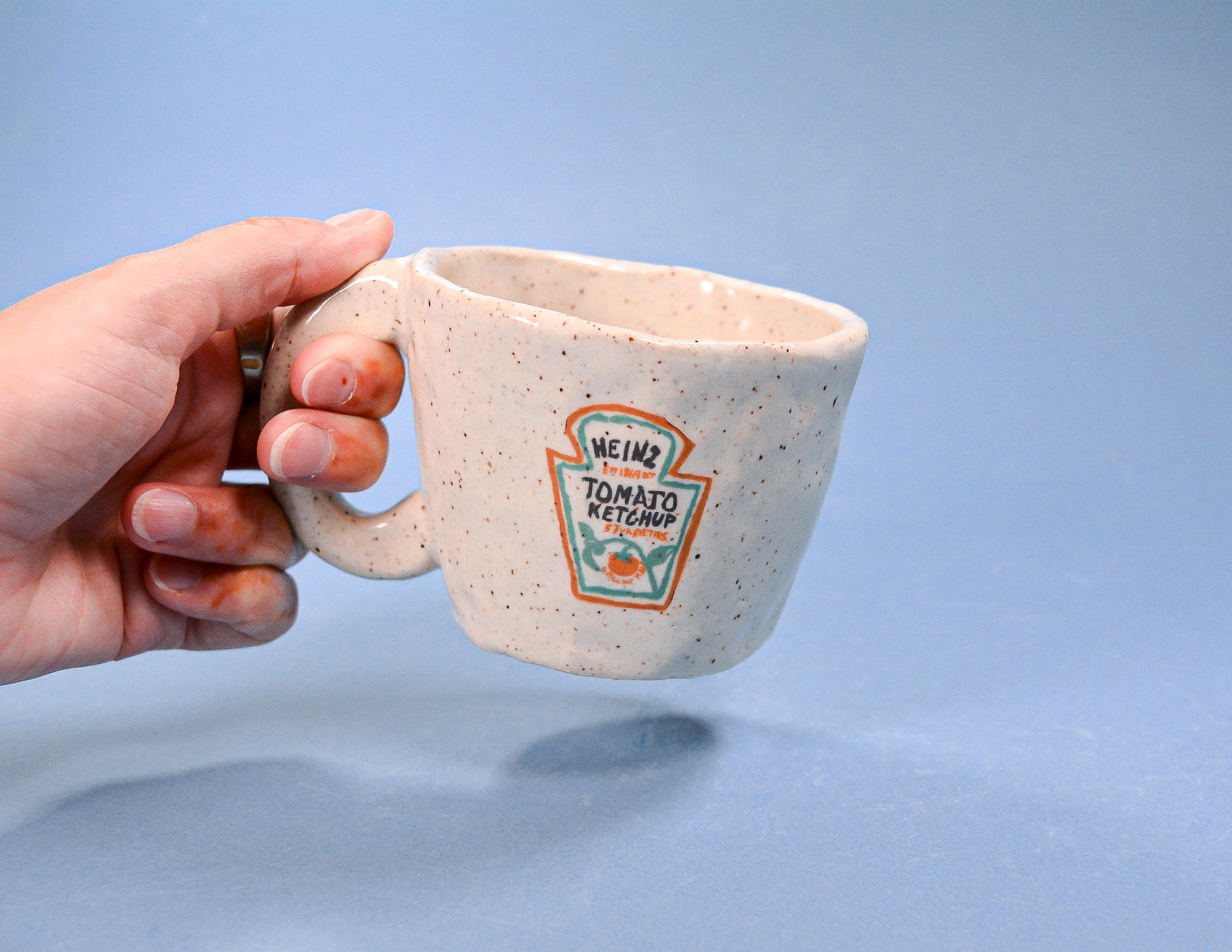 Hand holding a ceramic cup with Heinz ketchup logo against a blue background