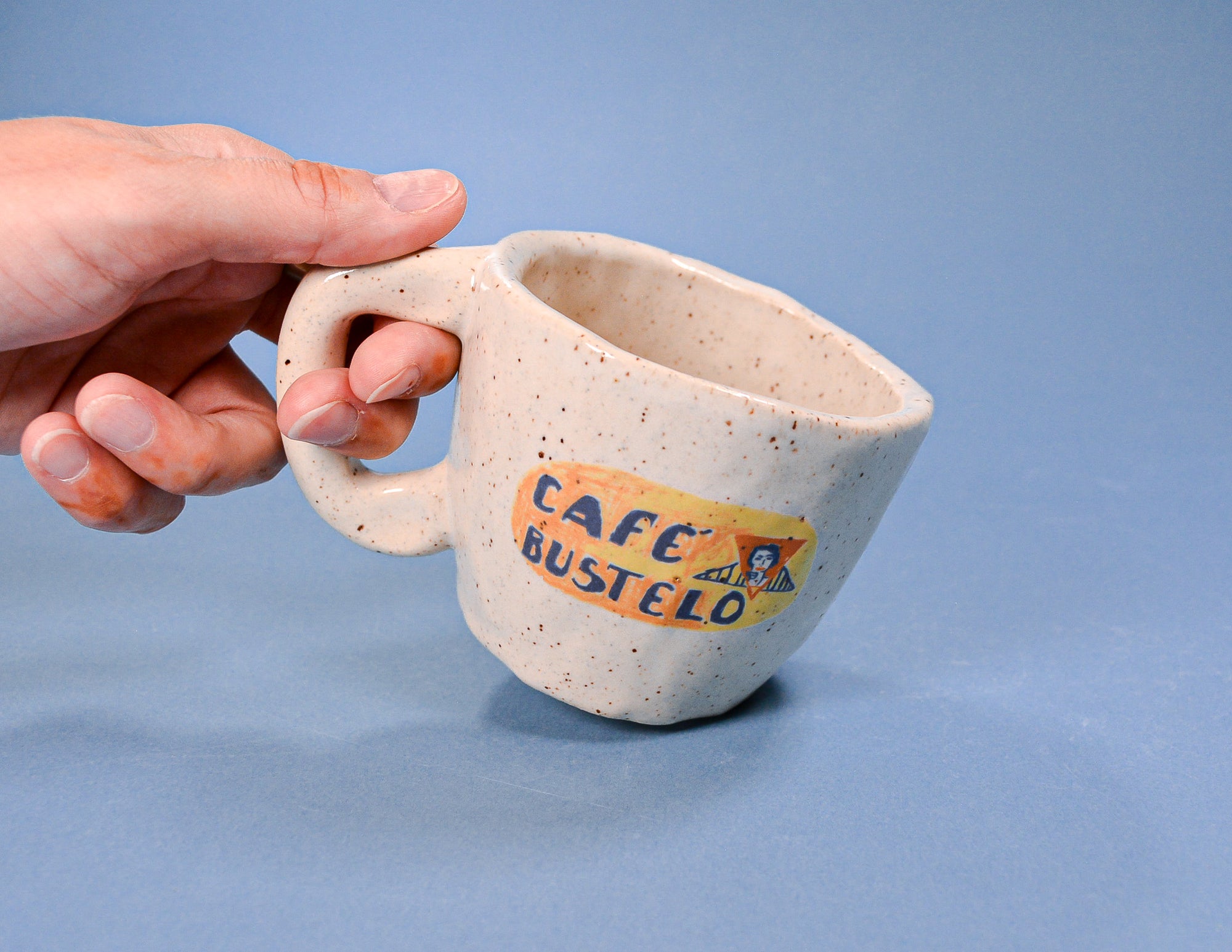 Hand holding a speckled ceramic mug with Cafe Bustelo logo on a blue background