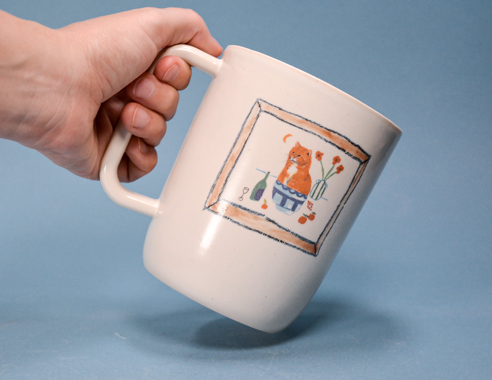 A hand holding a cream-colored ceramic mug with a hand-painted illustration of an orange cat in a blue pot on a blue background.