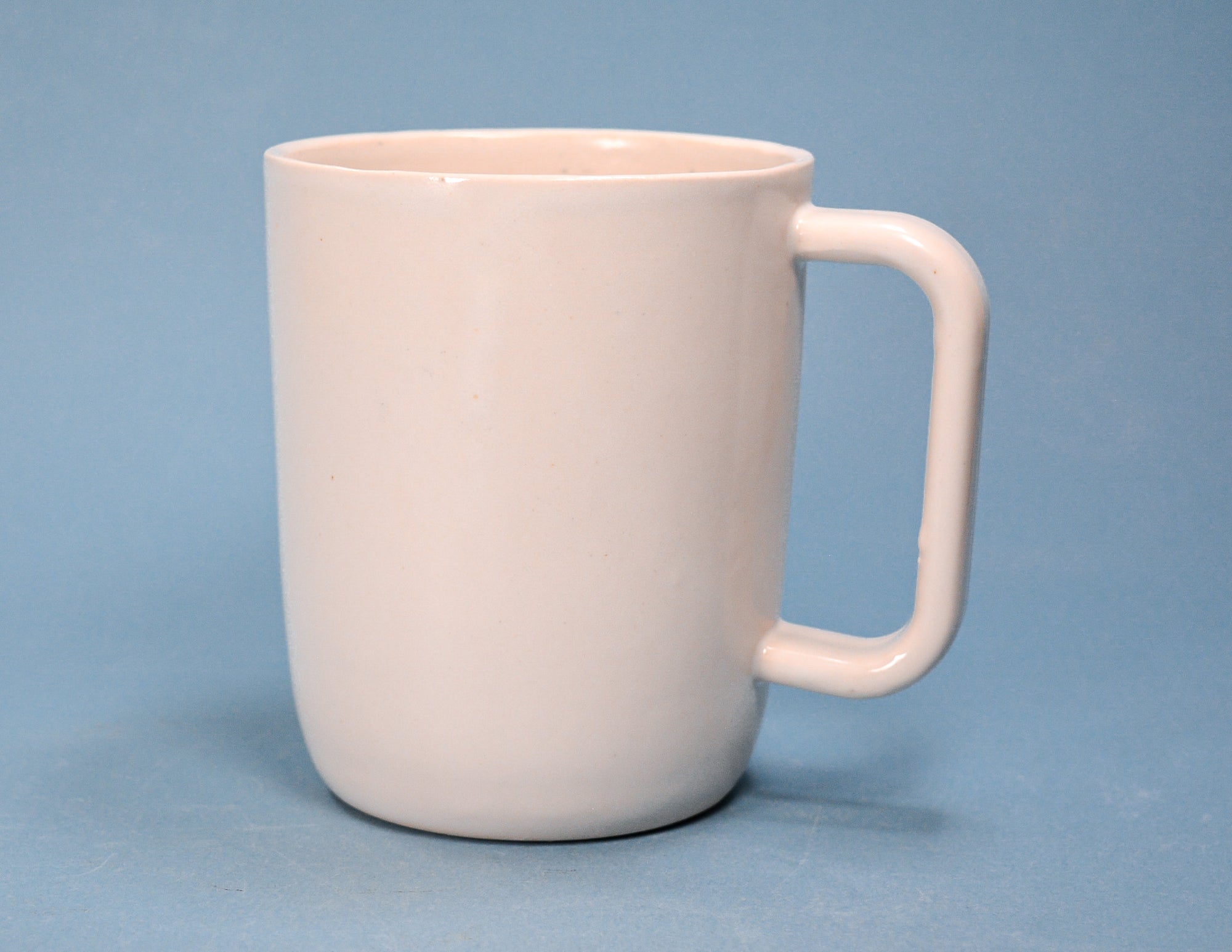 A classic ceramic coffee mug with a rounded body and a square-profiled handle, shown in a solid pale pink color against a solid blue background.