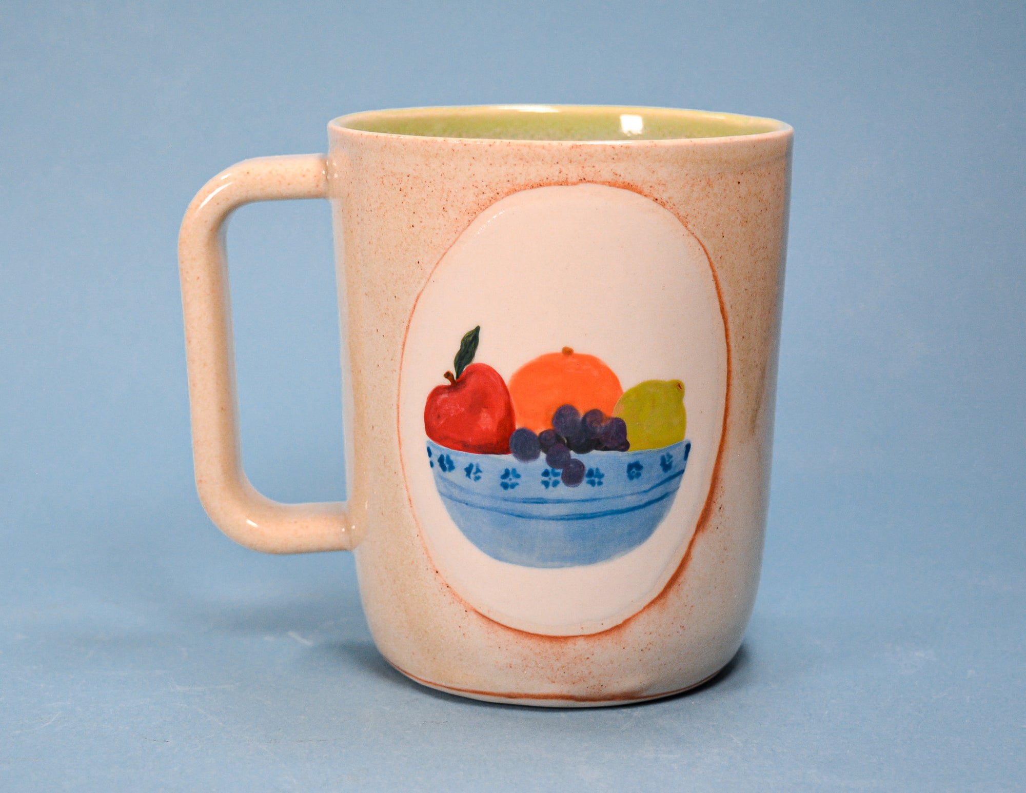 A speckled beige ceramic mug with a handle, featuring a hand-painted fruit illustration on the front, set against a solid blue background.