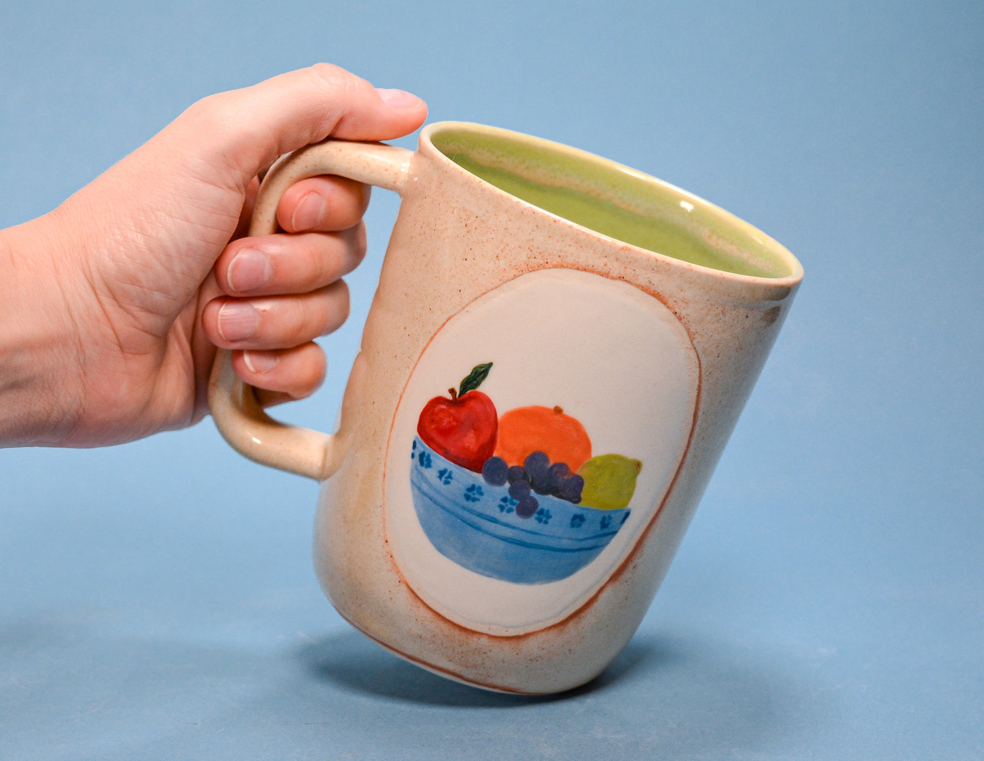 A ceramic mug with a hand-held handle, featuring a hand-painted fruit illustration on the front. The mug has a speckled beige exterior, a light green interior, and is held by a hand against a solid blue background.