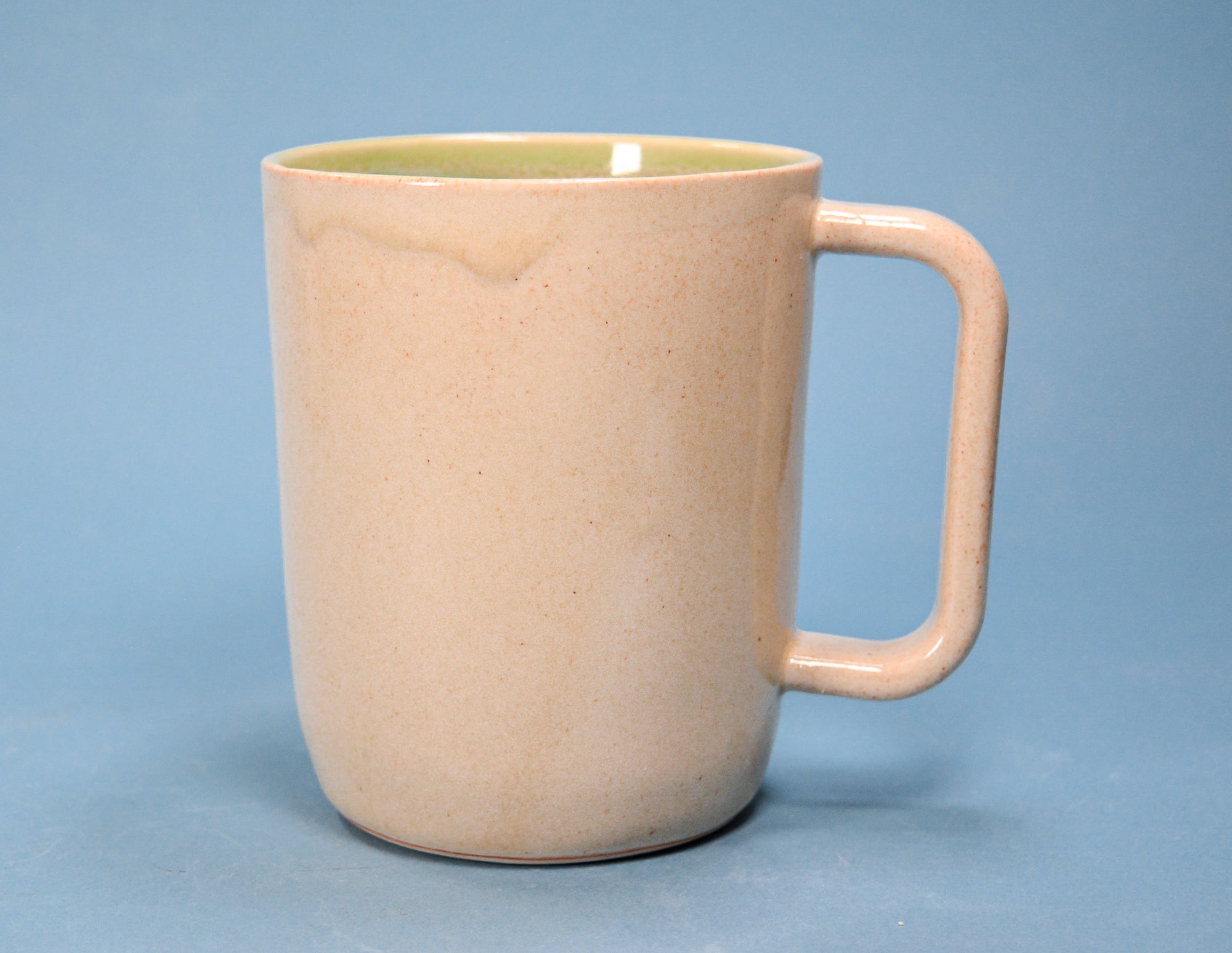 A speckled ceramic mug with a warm beige exterior and a light green interior, featuring a thick integrated handle, set against a solid blue background.