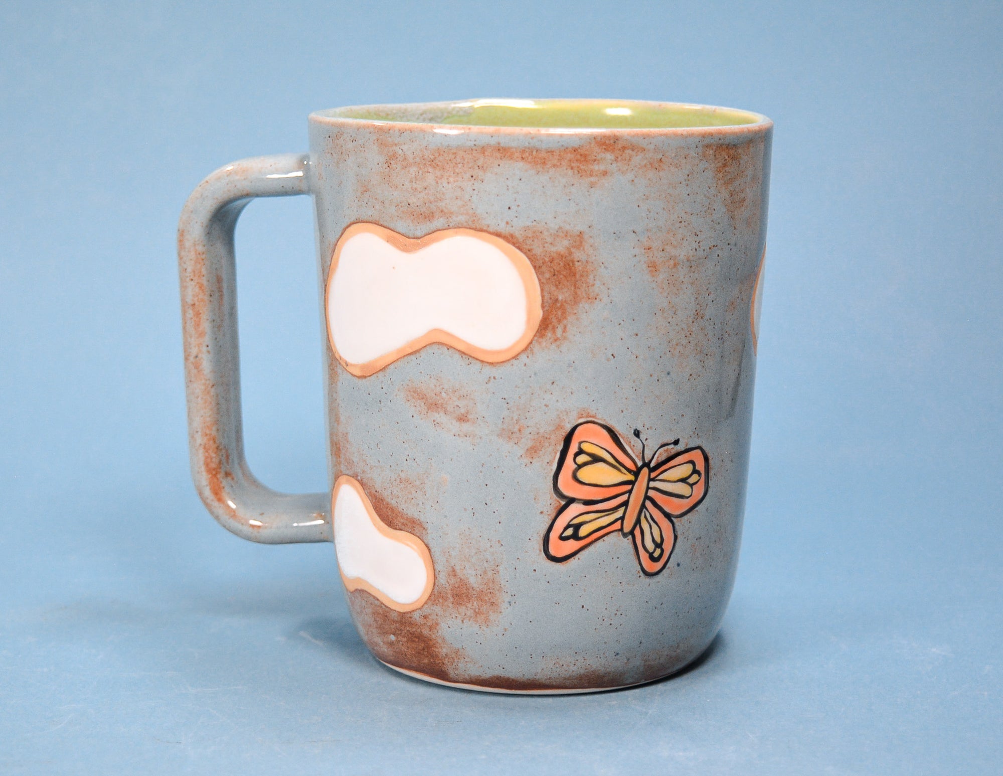 A ceramic mug with a speckled light blue-gray exterior, brown distressed patches, a painted orange butterfly, and two white cloud-like shapes, shown against a solid blue background.