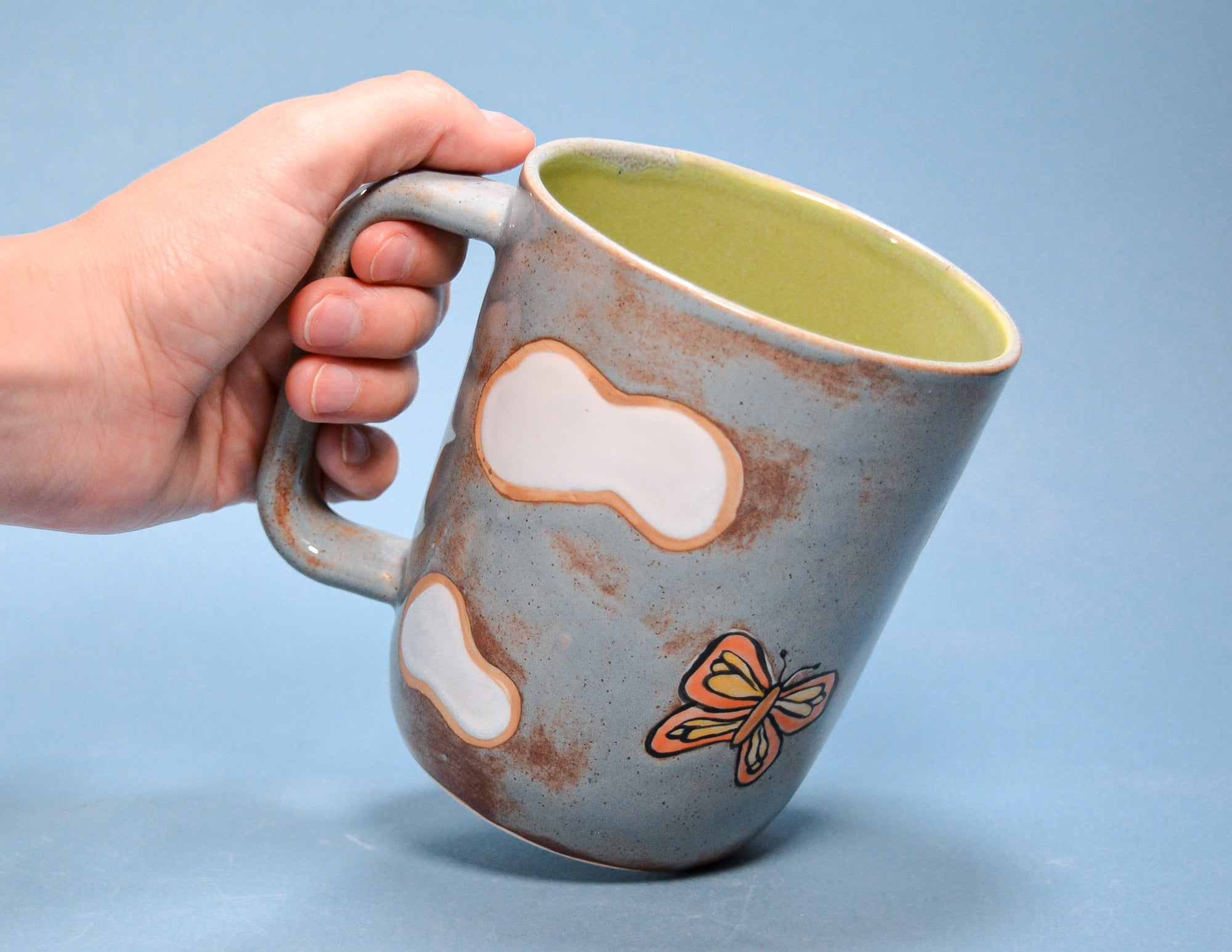 A hand holding a handmade ceramic mug with a distressed slate blue exterior, white cow-print style patches, a small orange butterfly, and a light green interior, set against a solid blue background.