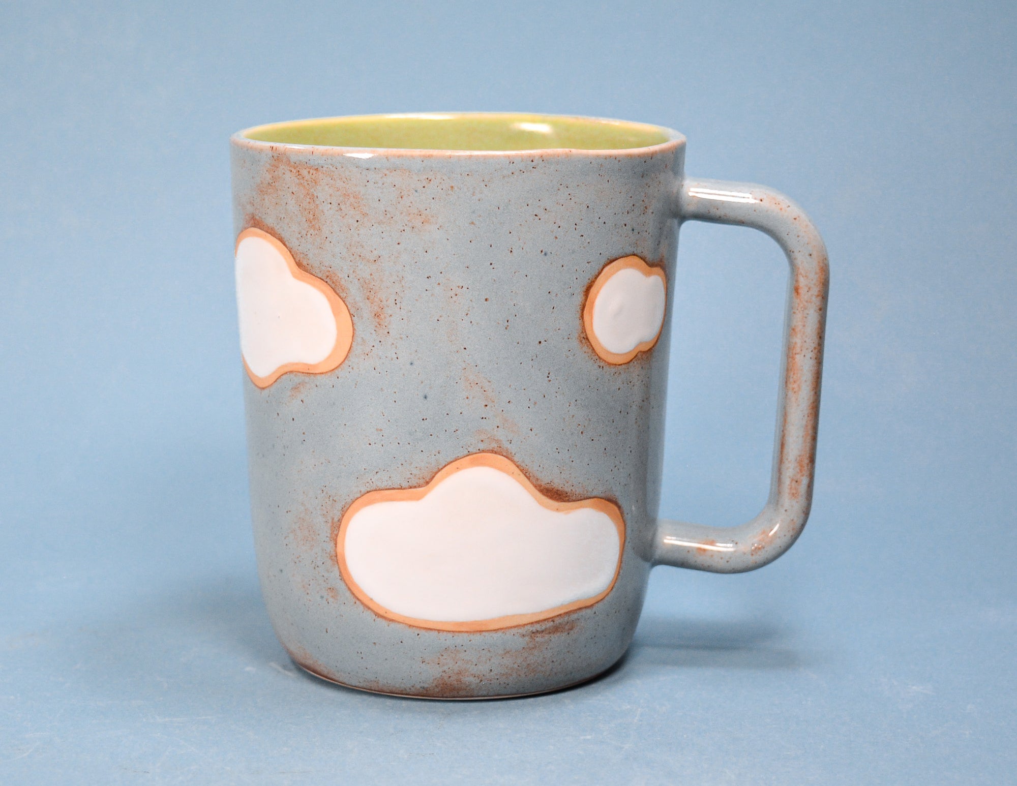 A speckled gray ceramic mug with a handle, featuring hand-painted white cloud shapes outlined in orange, with a light yellow interior, set against a solid blue background.