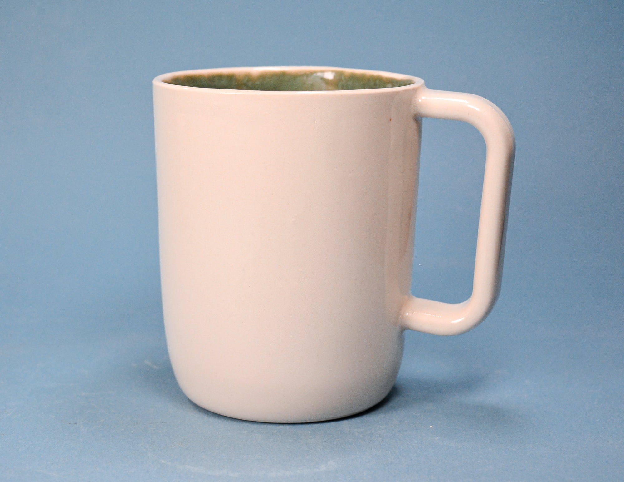 A ceramic mug with a matte off-white exterior and a textured olive-green interior, shown against a solid blue background.