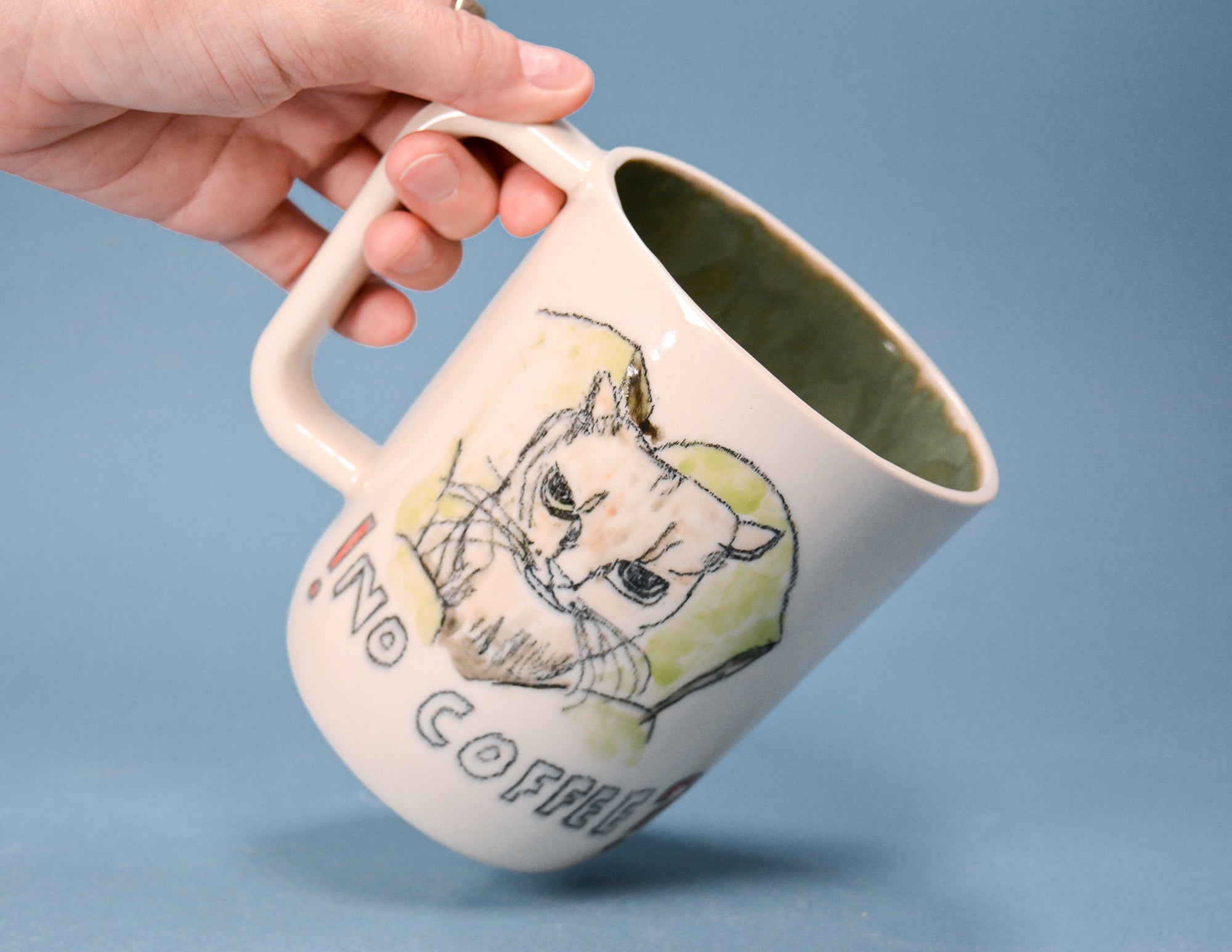A hand holds a ceramic coffee mug with a hand-drawn illustration of a grumpy cat and the text 'NO COFFEE' against a solid blue background.