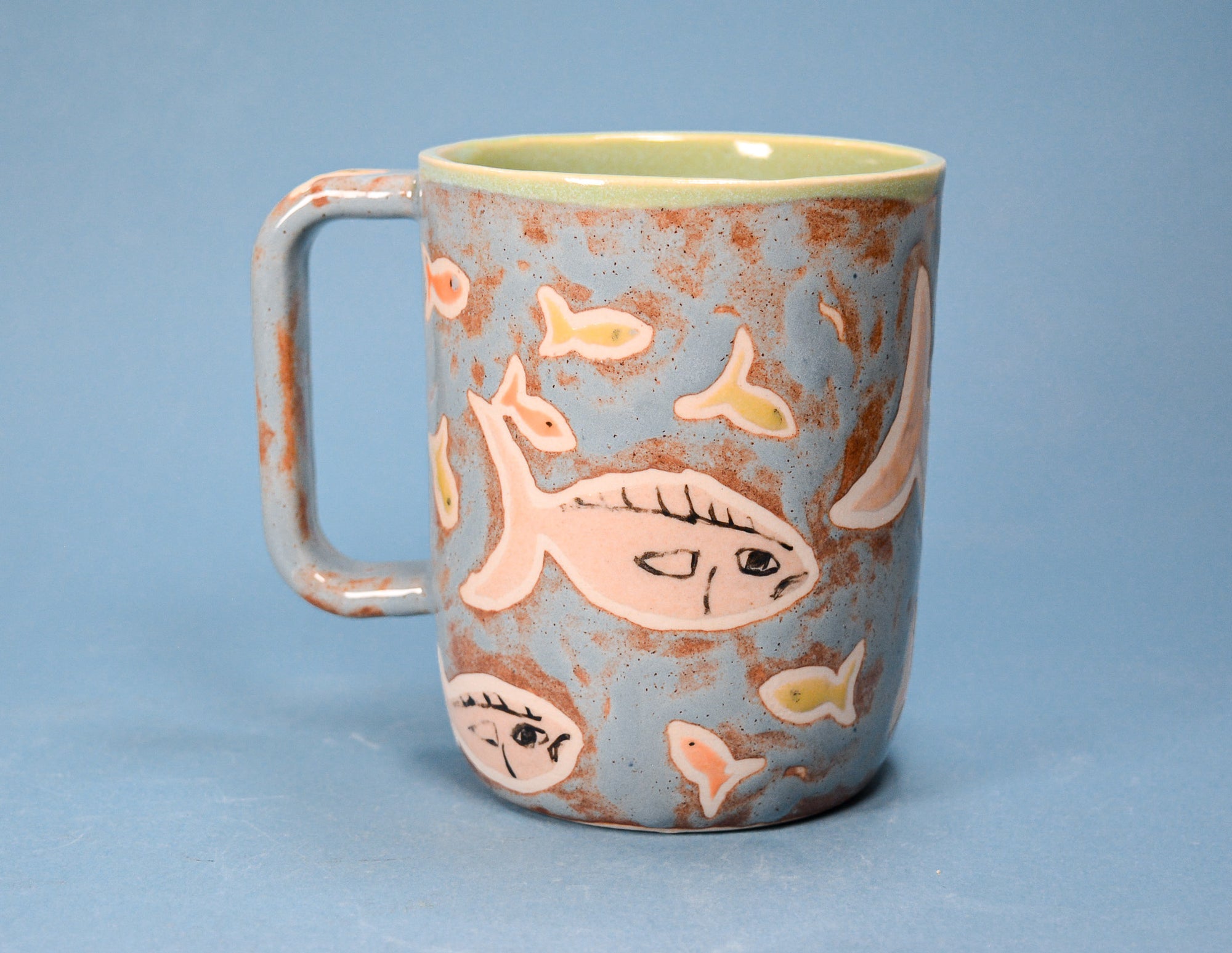 A ceramic mug with a speckled light blue exterior, decorated with hand-painted pink and yellow fish, and a solid pale green interior, set against a solid blue background.