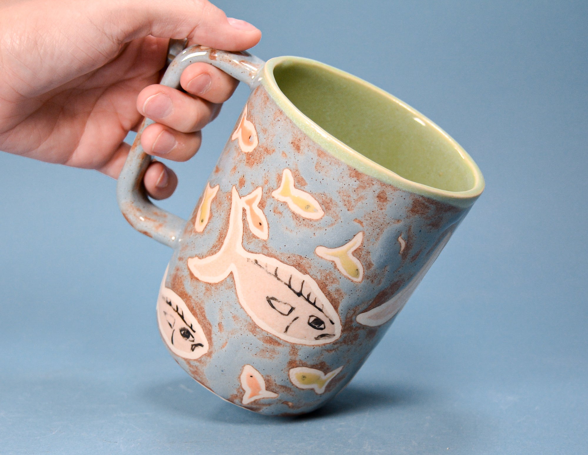A hand holding a handmade ceramic mug with a blue-gray exterior decorated with hand-painted fish in pink, yellow, and black, featuring a light green glazed interior, set against a solid blue background.