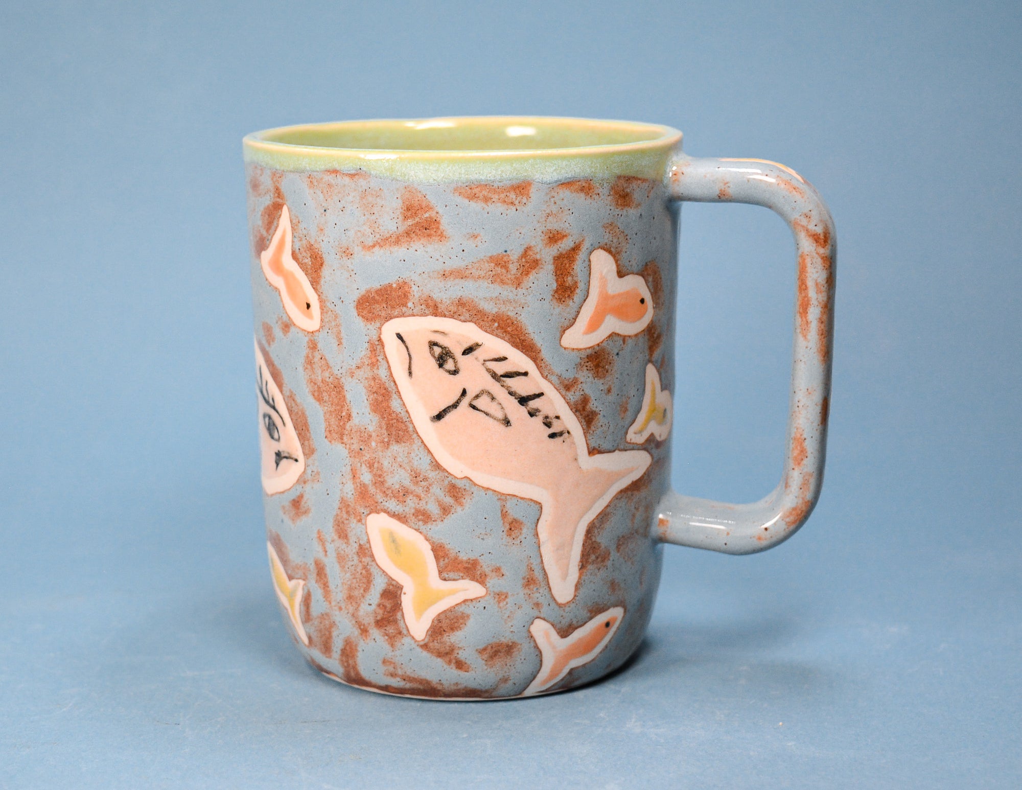 A ceramic mug with a speckled light blue and terracotta exterior, decorated with hand-painted pink fish, and a light green glazed interior, set against a solid blue background.