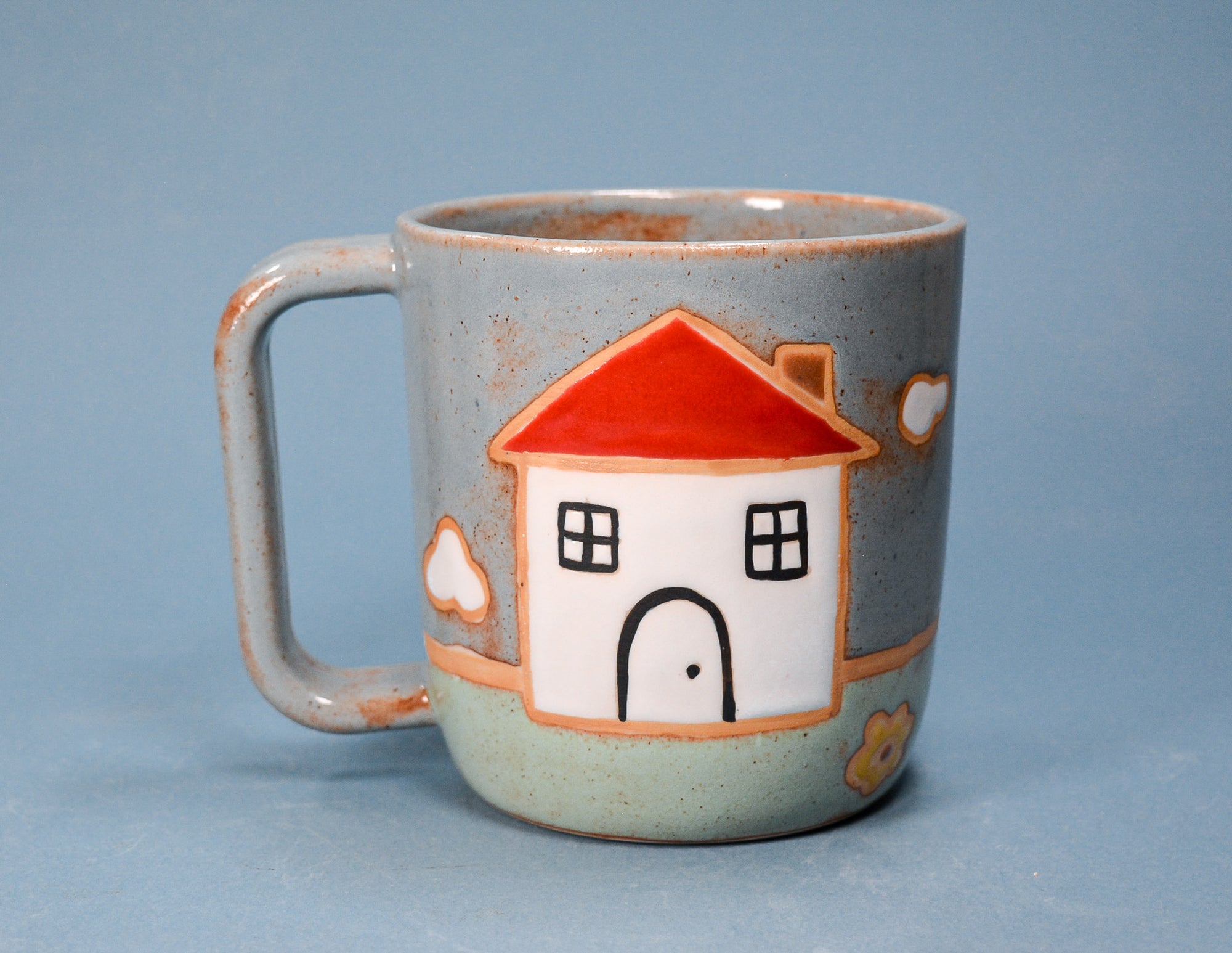 A speckled ceramic mug with a handle, featuring a hand-painted house design with a red roof, white body, and black outlines, set against a solid blue background.