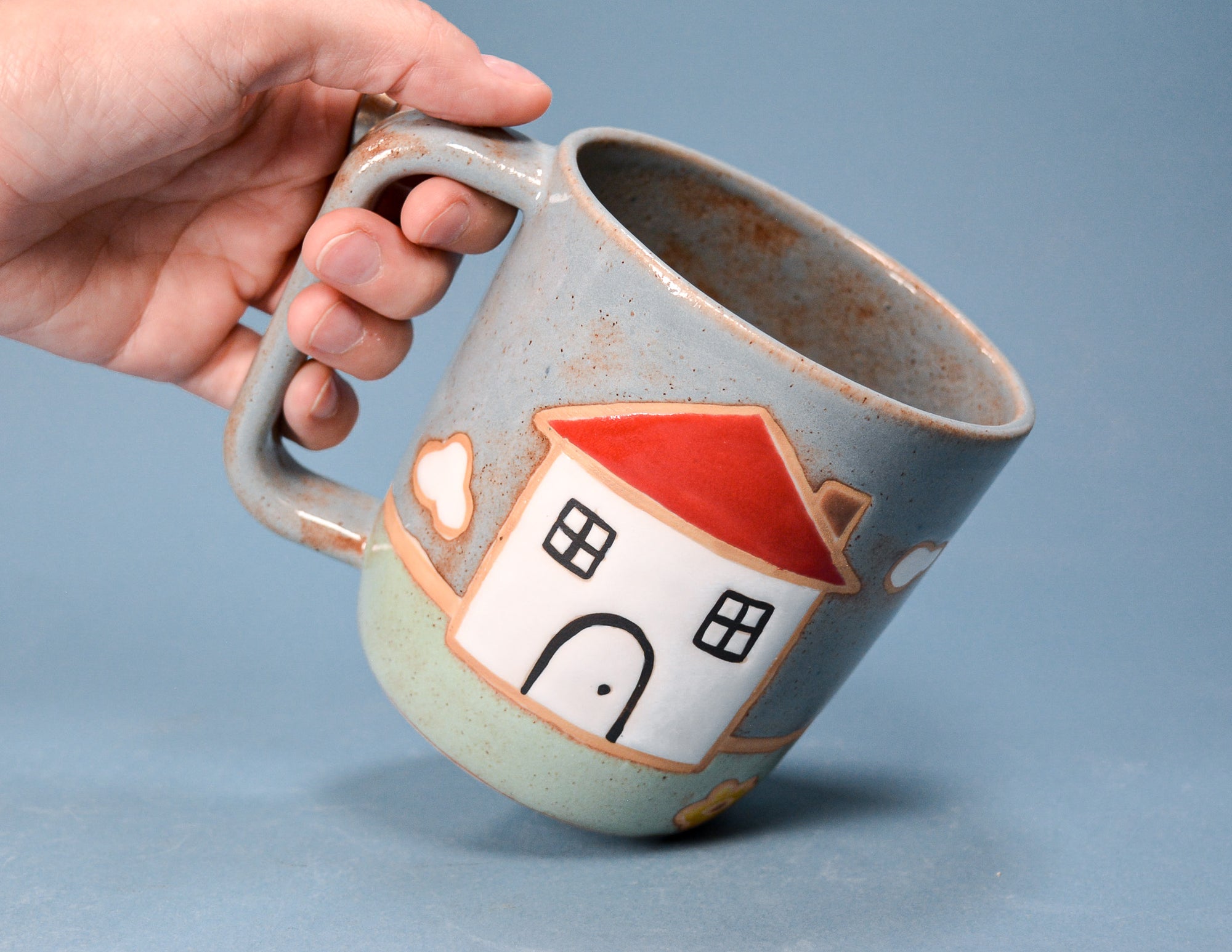 A hand holding a handmade ceramic mug with a handle. The mug has a speckled light blue-gray glaze, a white house illustration with a red roof, and a small white heart on the side, set against a solid blue background.
