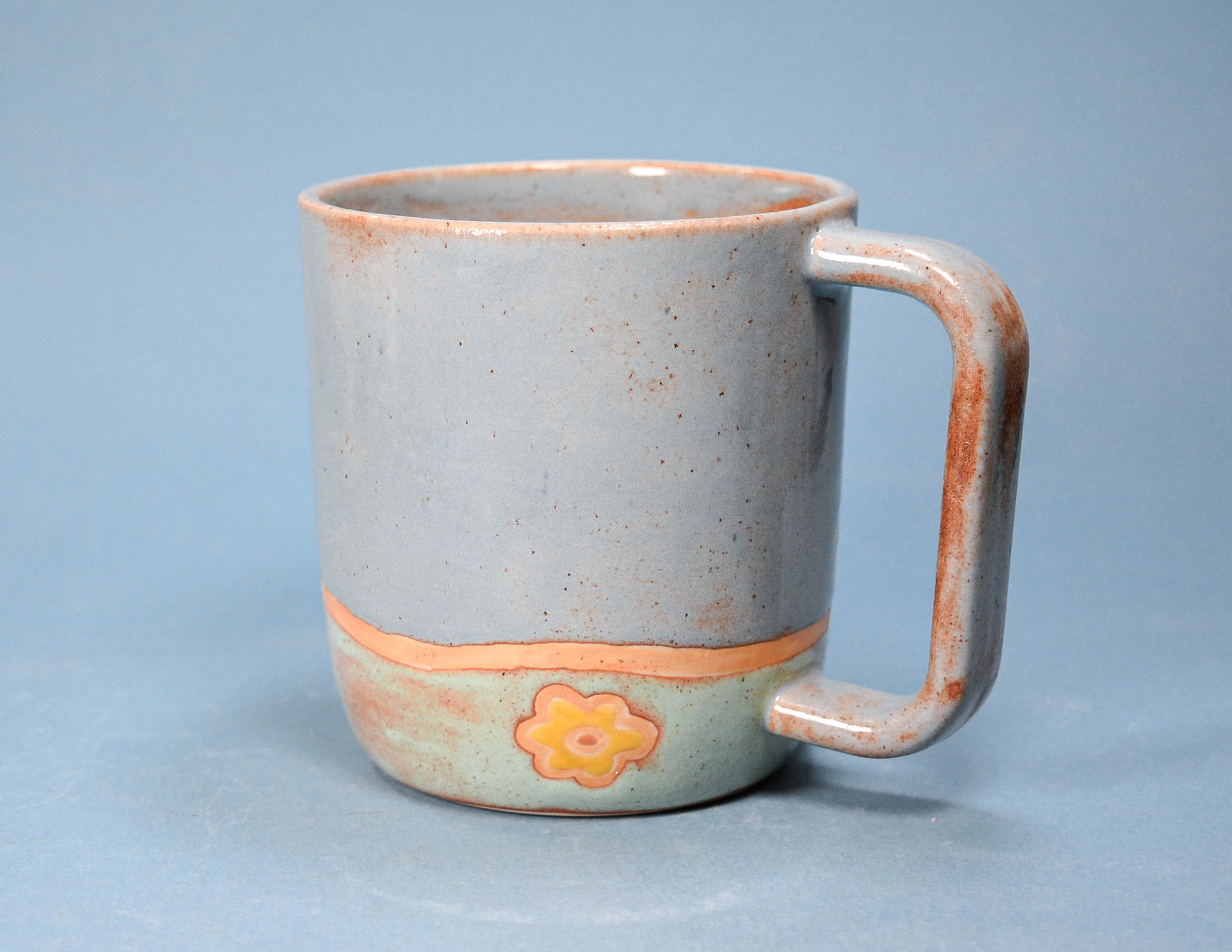 A handmade ceramic mug with a speckled light gray-blue glaze, a terracotta orange band around the lower half, and a small orange flower motif. The mug features a thick, integrated handle and is photographed against a solid light blue-gray background.