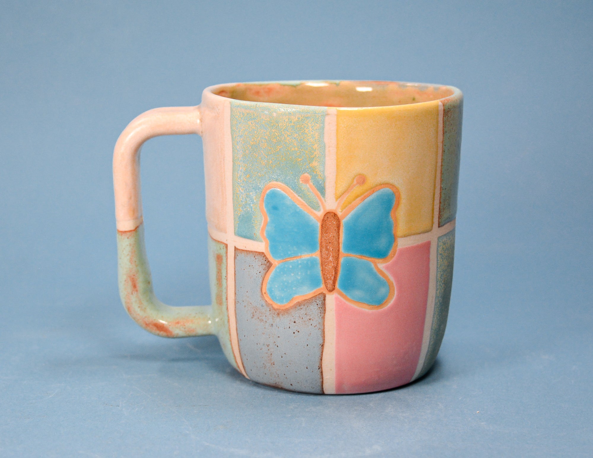 A ceramic mug with a pastel patchwork design featuring pink, yellow, blue, and gray sections, decorated with a blue butterfly motif in the center, set against a solid blue background.