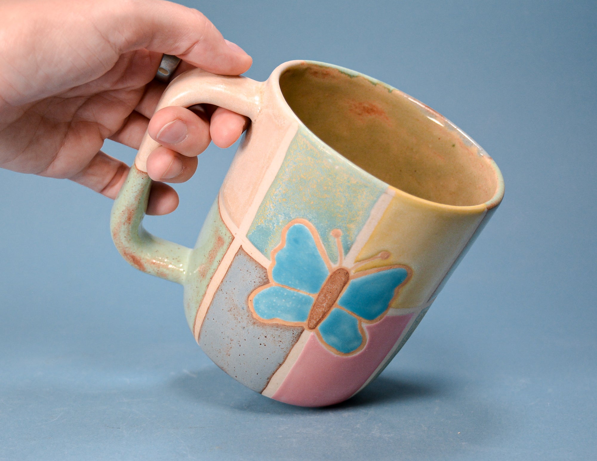A handmade ceramic mug with a pastel multicolor glaze and a blue butterfly design, held by a hand against a solid blue background.