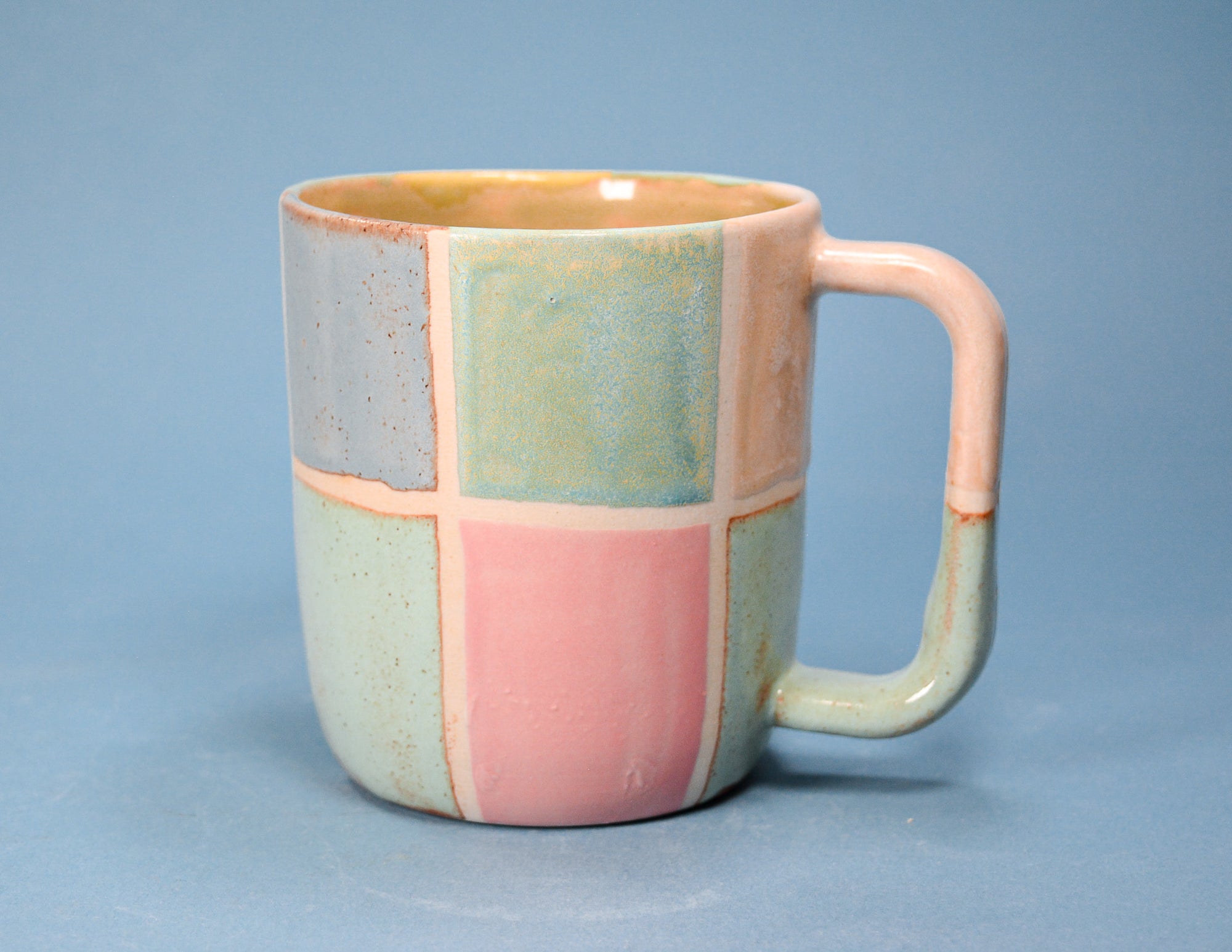 A ceramic mug with a patchwork design in pastel shades of pink, light blue, and sage green, featuring a warm orange-brown glaze on the interior and handle, set against a solid blue background.