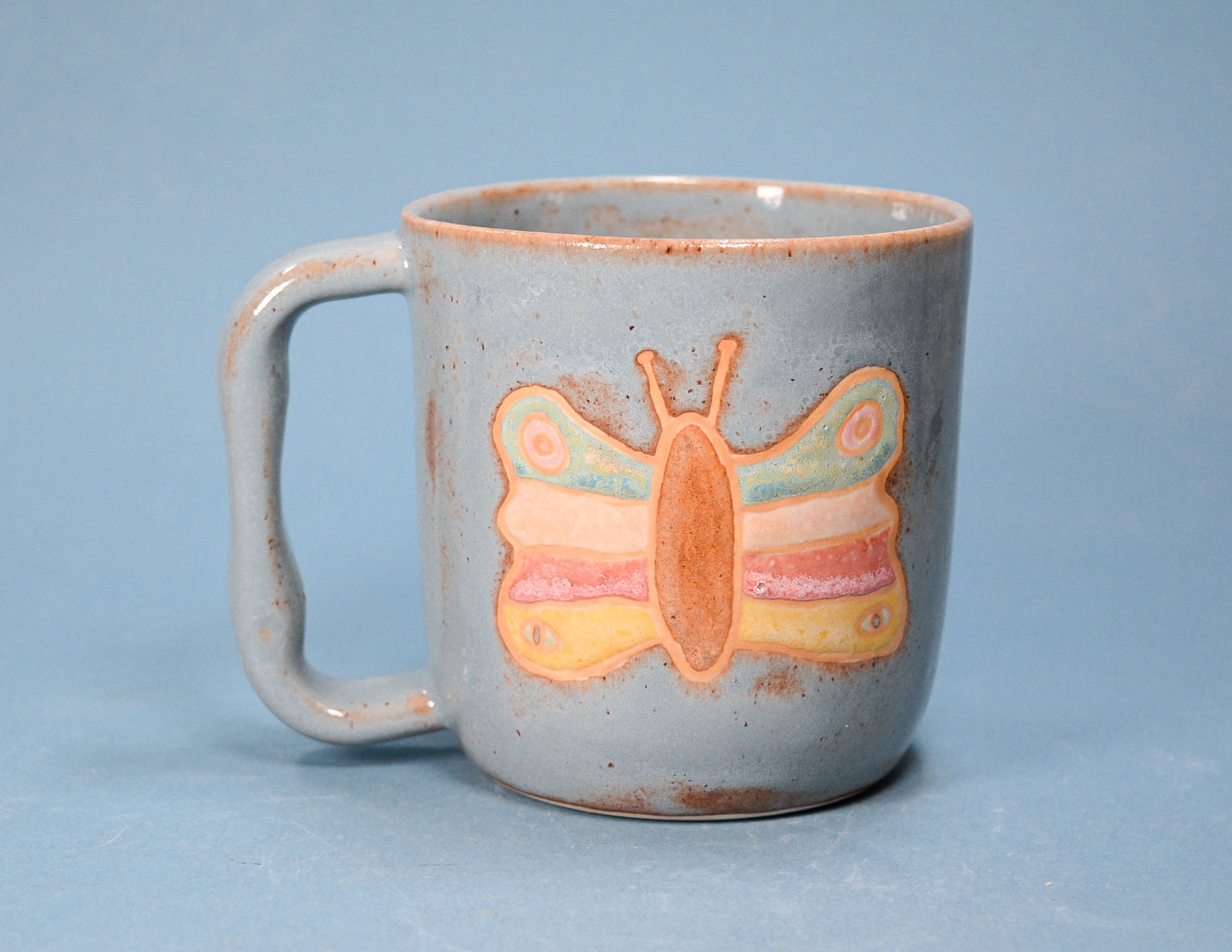 A speckled light gray ceramic mug with a handle, featuring a painted butterfly design in pastel orange, yellow, pink, and light blue on the front, set against a solid blue background.