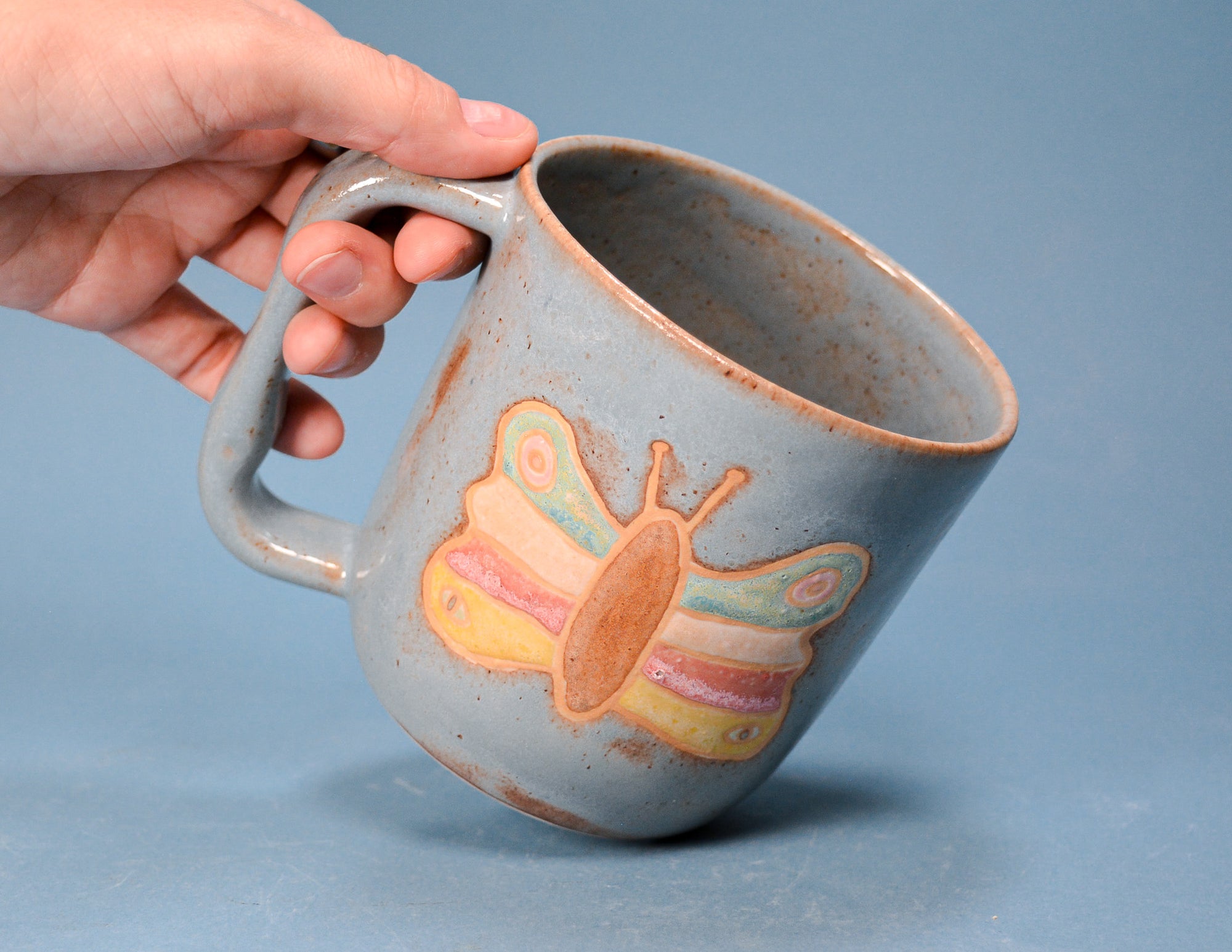 A hand holding a handmade ceramic mug with a light blue-gray glaze and a painted butterfly design in pastel yellow, pink, and green. The mug has a rustic, speckled brown rim and is set against a solid blue-gray background.