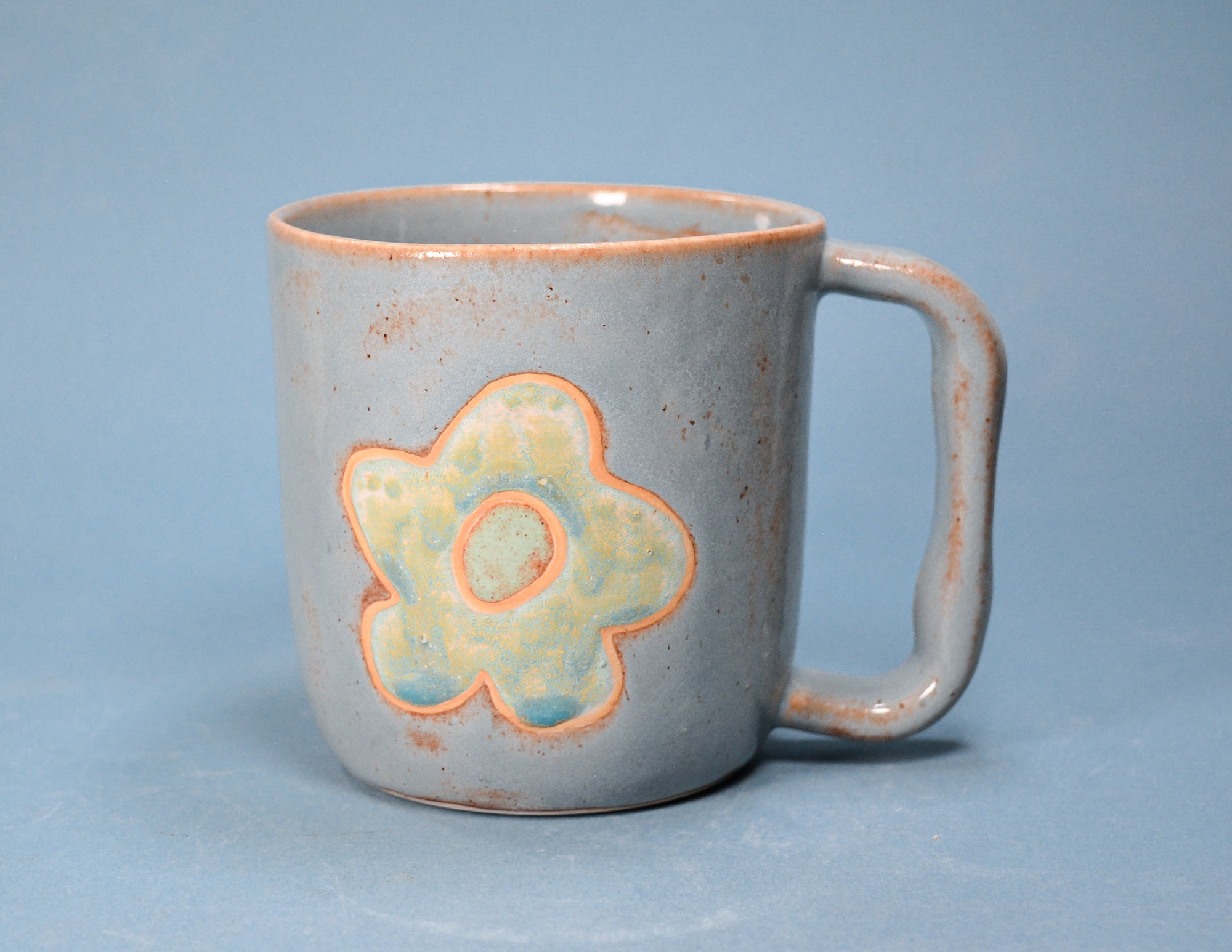 A handmade ceramic mug with a speckled light gray-blue glaze, featuring a stylized flower motif in pale yellow-green and light blue with a terracotta orange outline. The mug has a rustic, distressed finish on the rim and handle, and is set against a solid blue background.