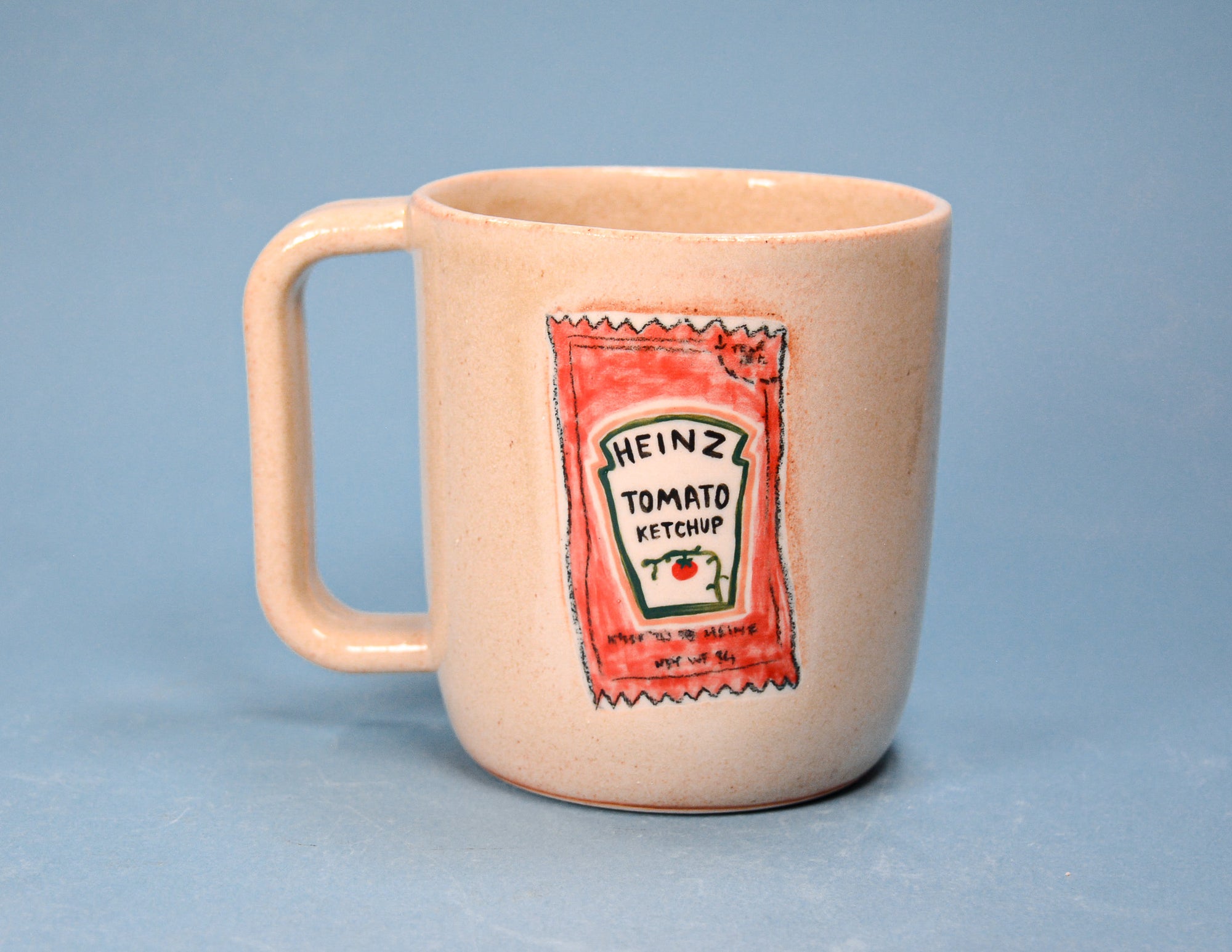 A speckled beige ceramic mug featuring a hand-drawn style red and white Heinz Tomato Ketchup bottle graphic on the front, set against a solid blue background.