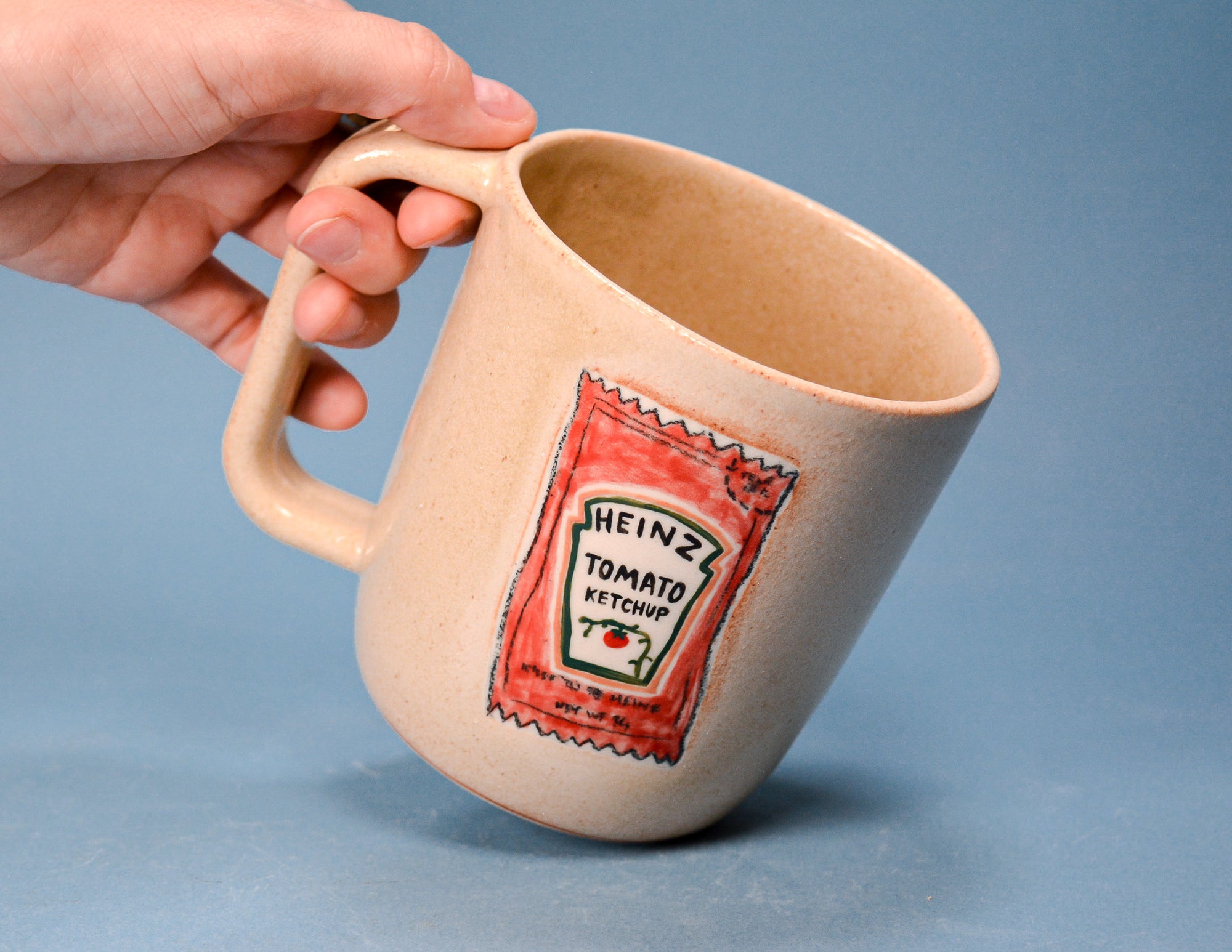 A hand holding a beige ceramic mug designed to look like a Heinz Tomato Ketchup bottle, set against a solid blue background.