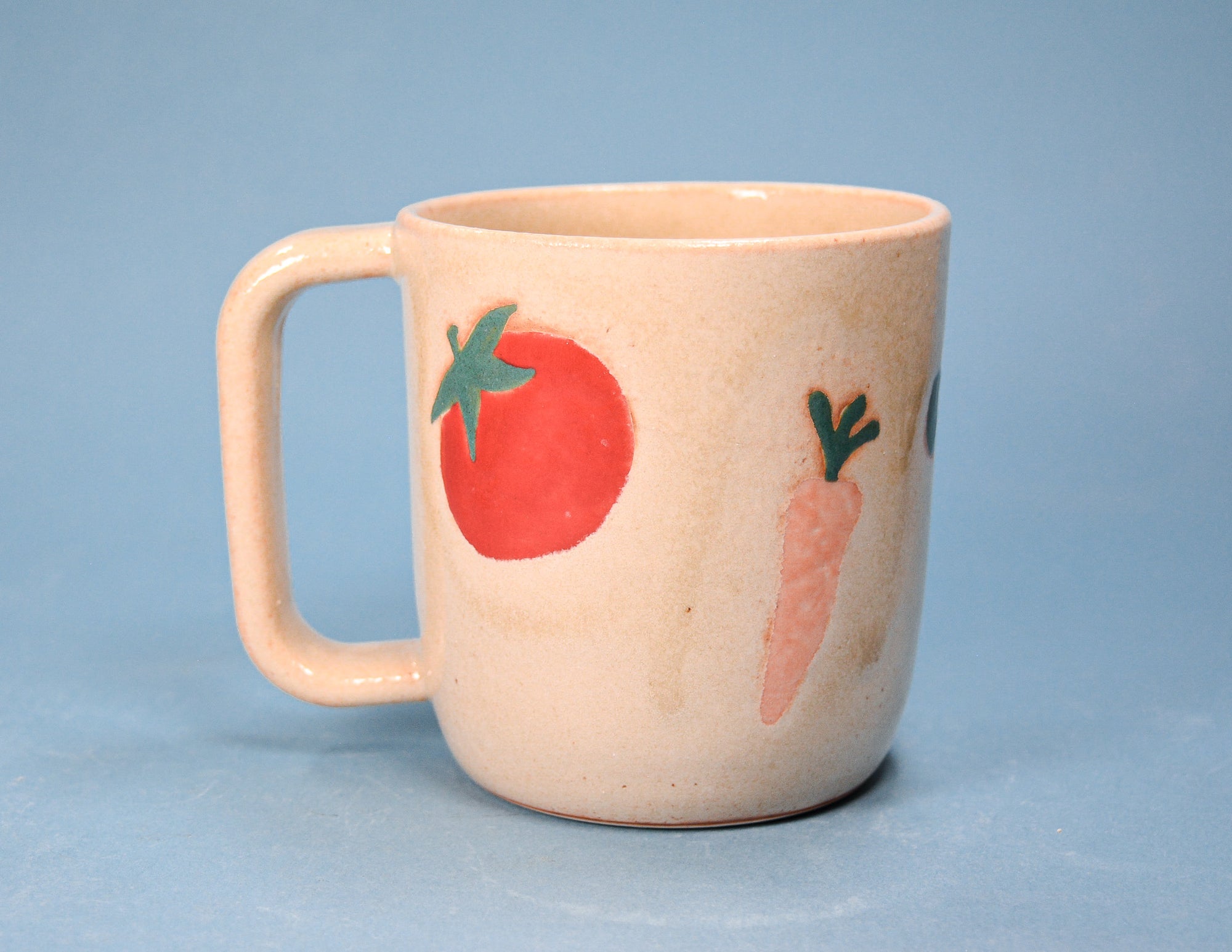 A beige ceramic mug with a rounded handle, decorated with a red tomato and a pink carrot painted on the side, set against a solid light blue background.