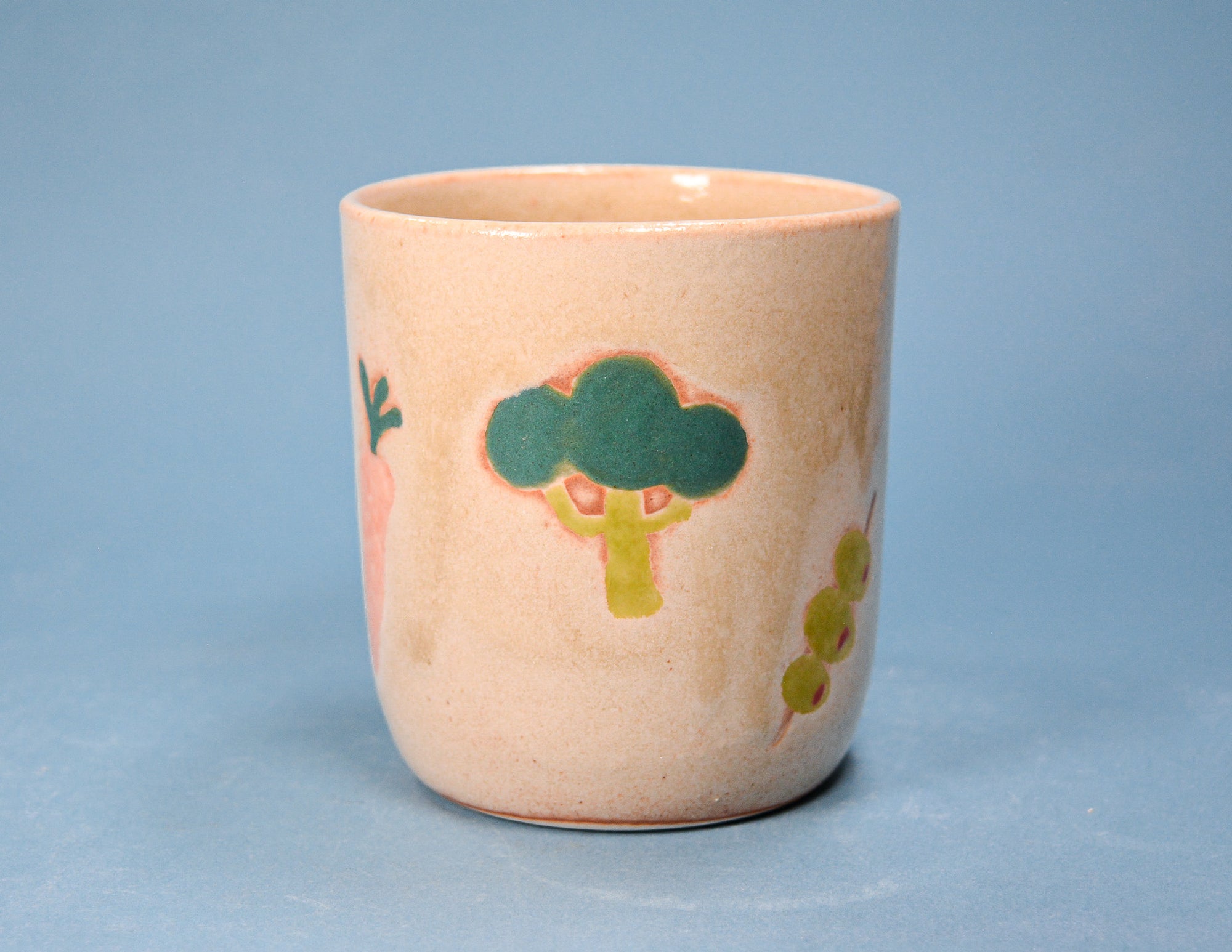 A small ceramic cup with a speckled beige-pink glaze, decorated with hand-painted vegetables including a green broccoli, pink carrot, and green peas, set against a solid blue background.