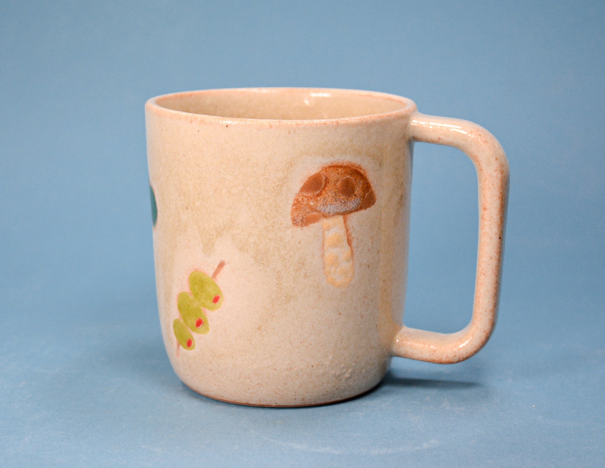 A speckled beige ceramic mug with a rounded handle, decorated with hand-painted illustrations of a brown mushroom and a green caterpillar on a soft blue background.