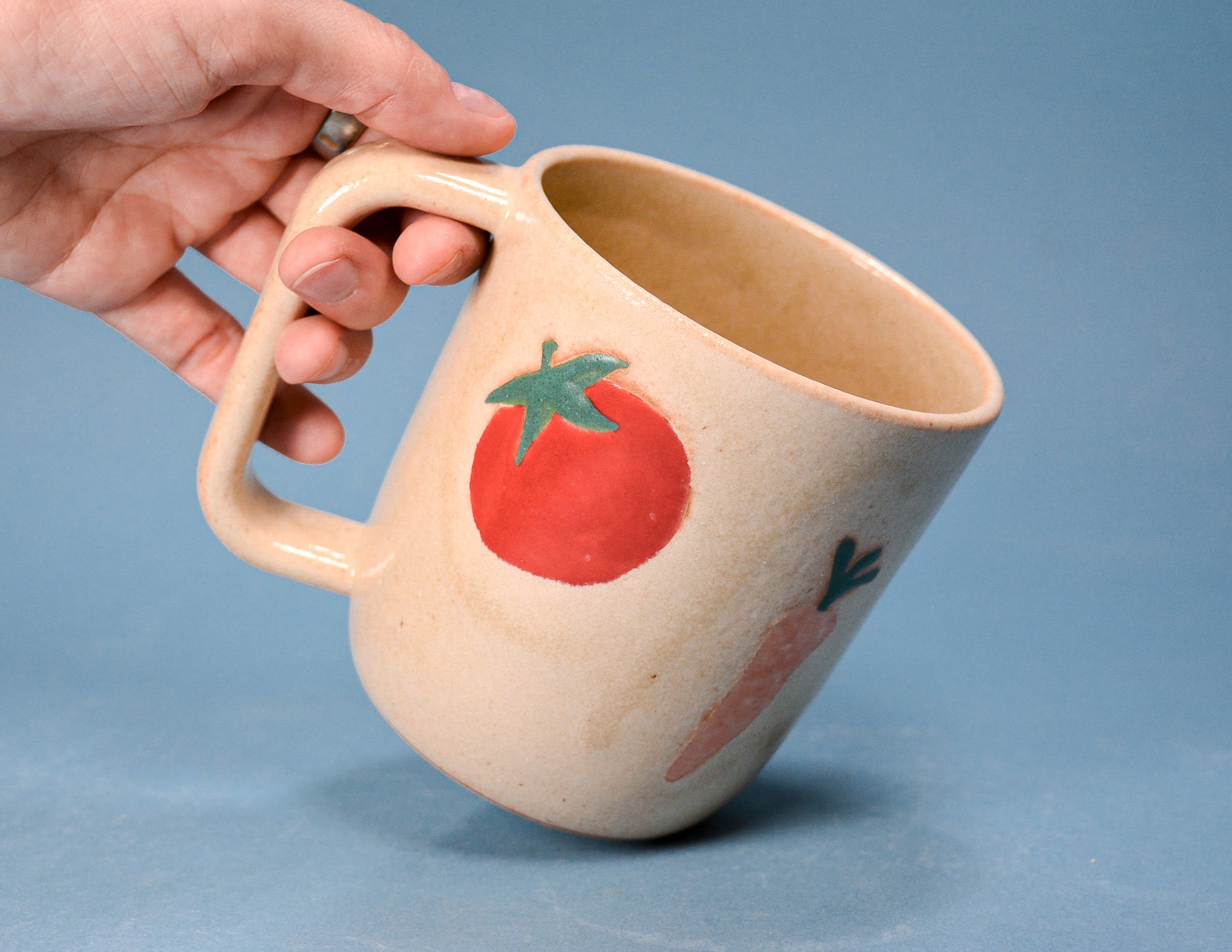A hand holding a beige ceramic mug with a painted red tomato and carrot design, set against a solid blue background.