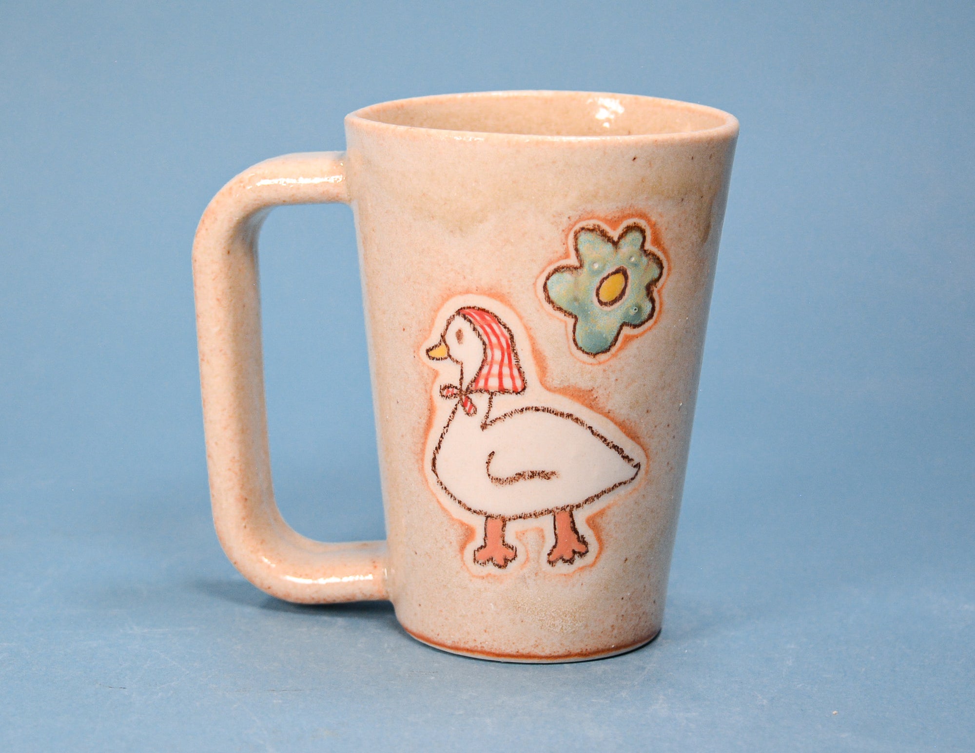 A speckled beige ceramic mug with a rounded handle, decorated with a white goose and a green flower motif on a solid blue background.