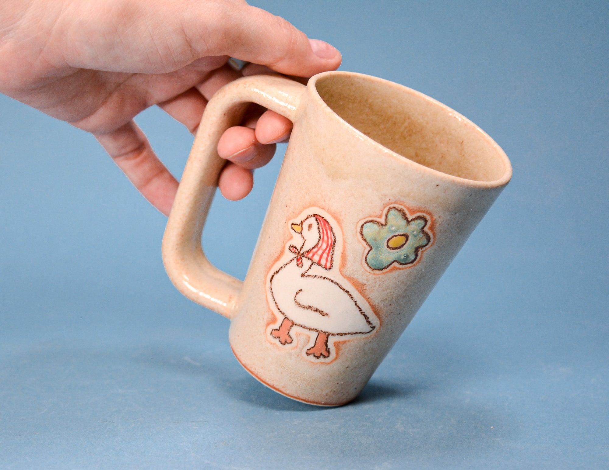 A hand holding a handmade ceramic mug with a handle. The mug has a warm beige glaze and is decorated with a white goose and a blue flower with orange outlines, set against a solid blue background.