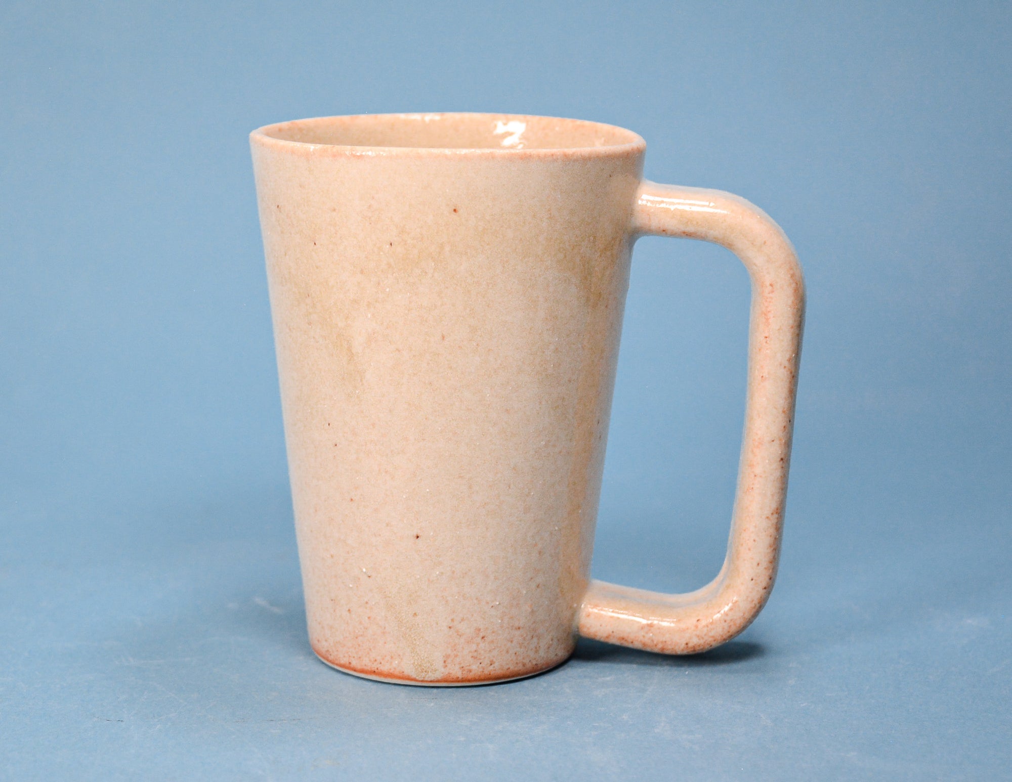 A handmade ceramic mug with a rounded handle and a speckled, matte finish in a light peach-beige color, set against a solid blue background.
