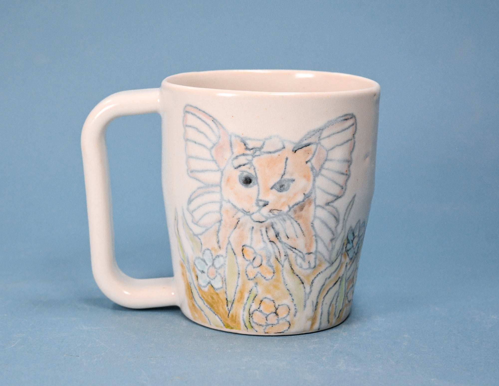 A white ceramic mug featuring a hand-painted illustration of a fairy cat with wings and yellow flowers, set against a solid blue background.