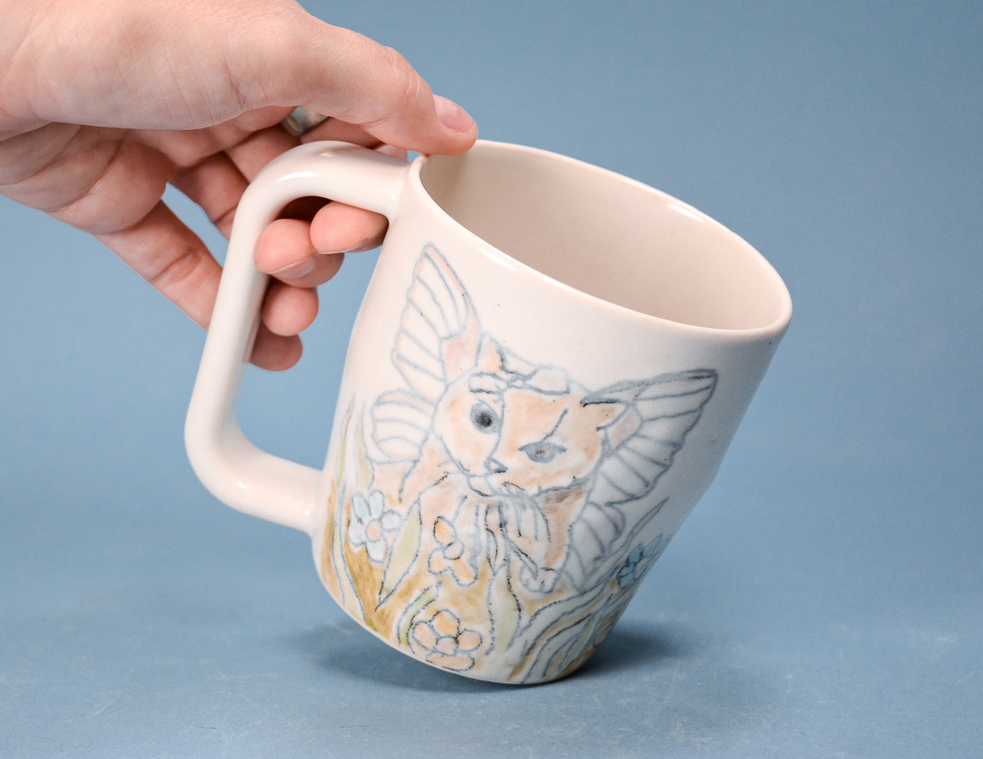 A hand holding a cream-colored ceramic mug with a detailed illustration of a winged fairy or angel cat in pastel orange, yellow, and blue-gray tones, set against a solid blue background.