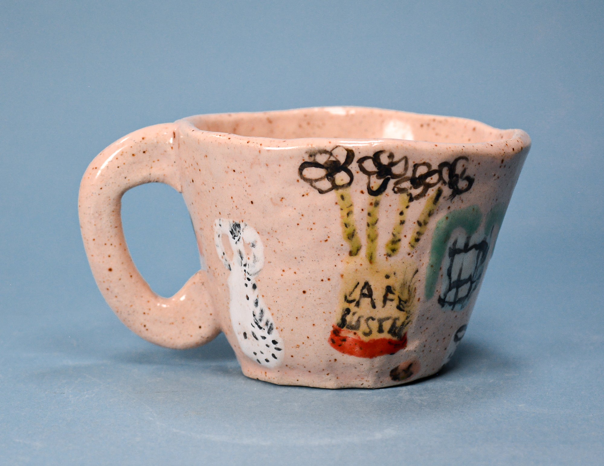 A handmade speckled ceramic coffee mug with a handle, decorated with hand-painted doodles and the words 'Cafe Mysterio' in yellow and red, set against a solid blue background.