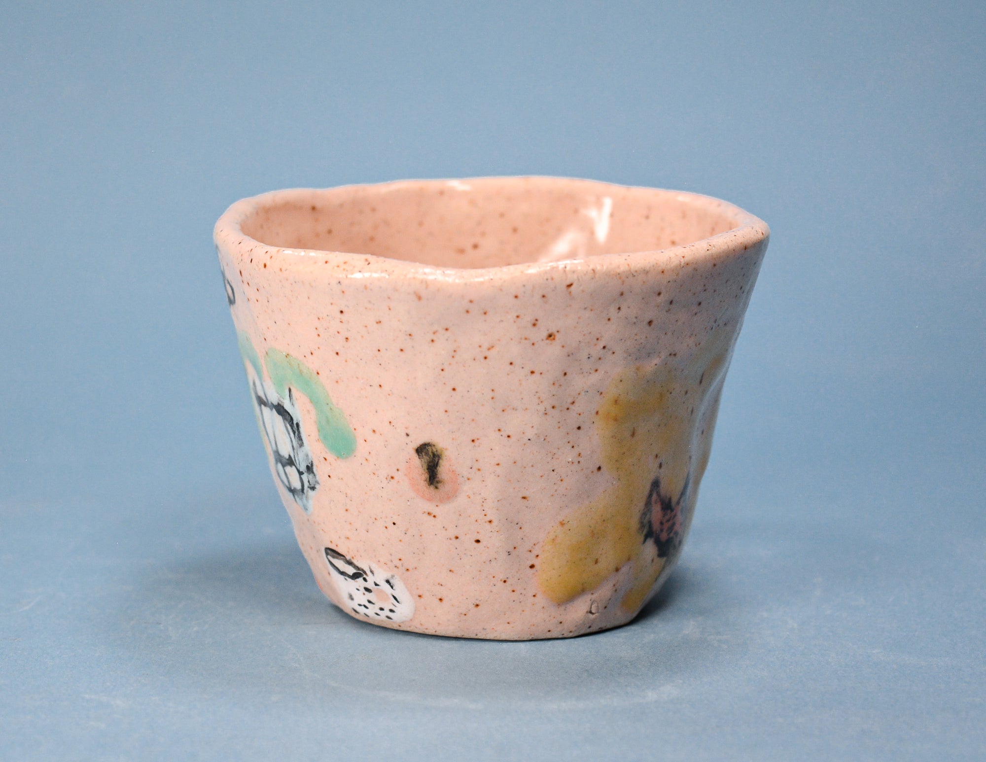 A handmade ceramic cup with a speckled pinkish-beige glaze, featuring hand-painted accents of yellow, green, and white, set against a solid blue-gray background.