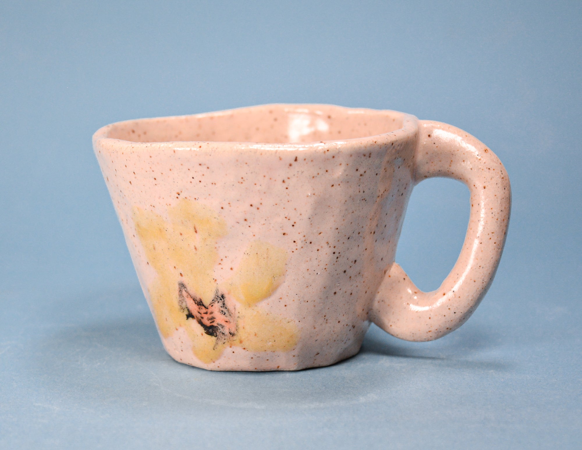 A handmade ceramic coffee cup with a light pink speckled glaze, featuring a yellow floral design on the lower left side, set against a solid blue background.