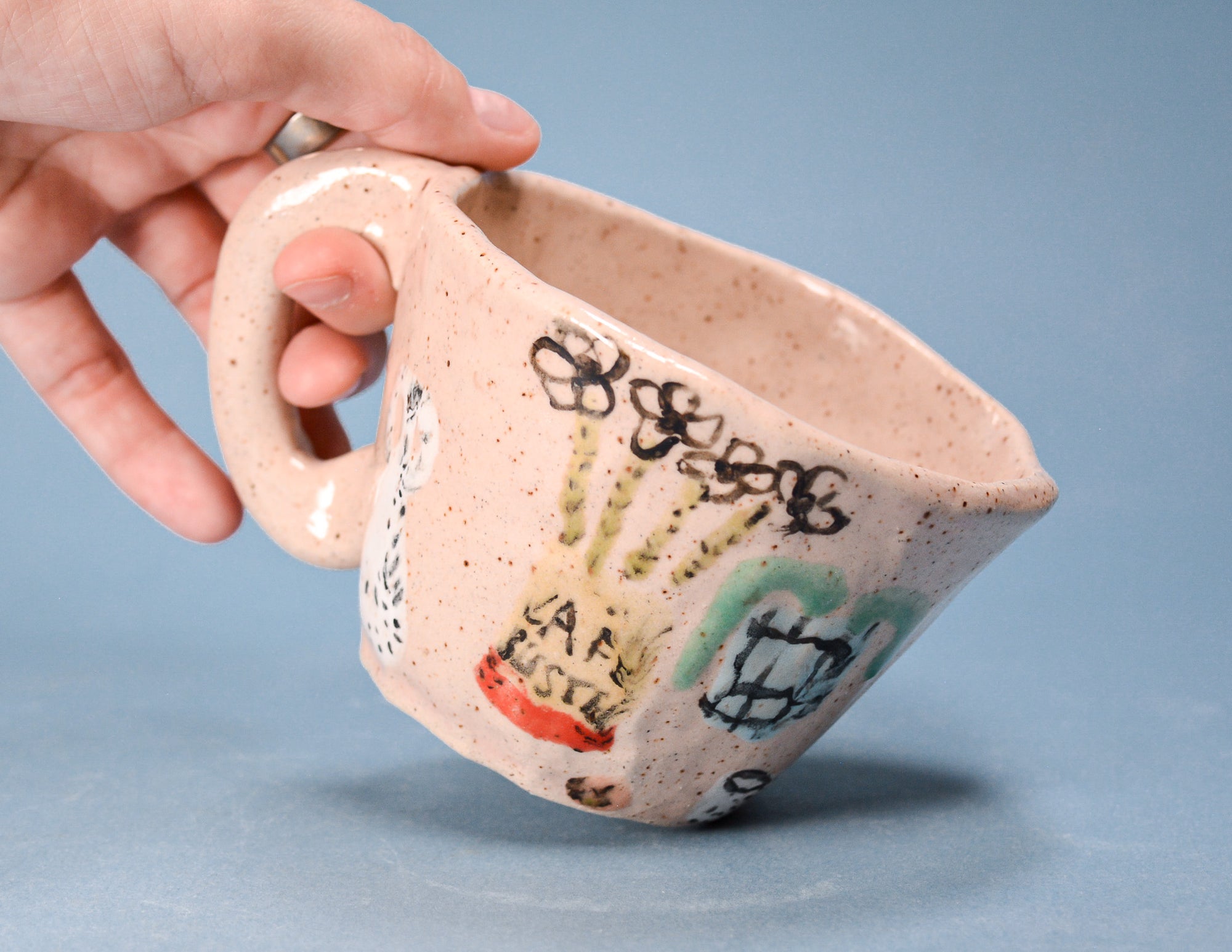 A hand holding a handmade ceramic mug with a speckled beige glaze and colorful hand-painted doodles, set against a solid blue background.
