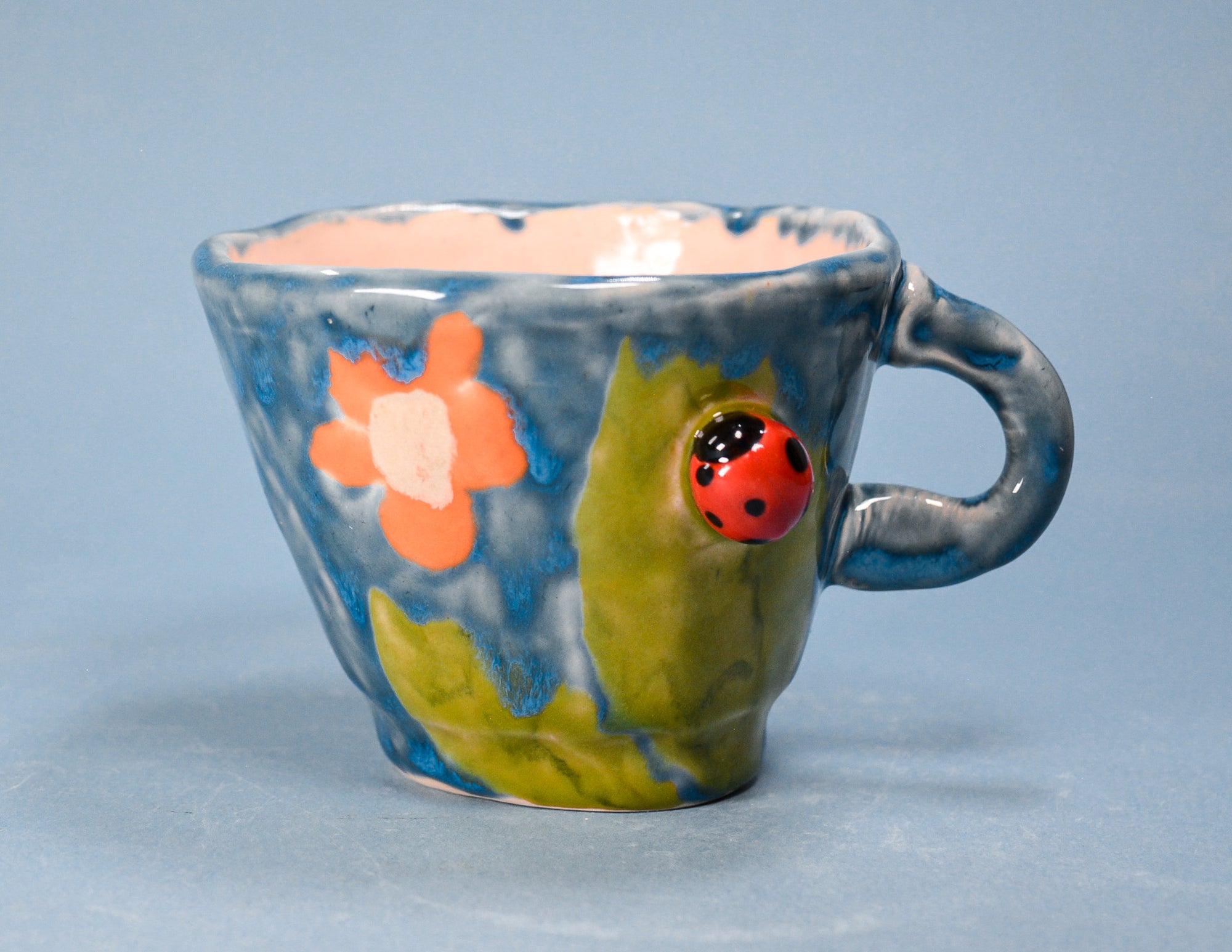 A handmade ceramic coffee cup with a blue and green marbled glaze, decorated with an orange flower, a red ladybug, and a small handle, set against a solid light blue background.