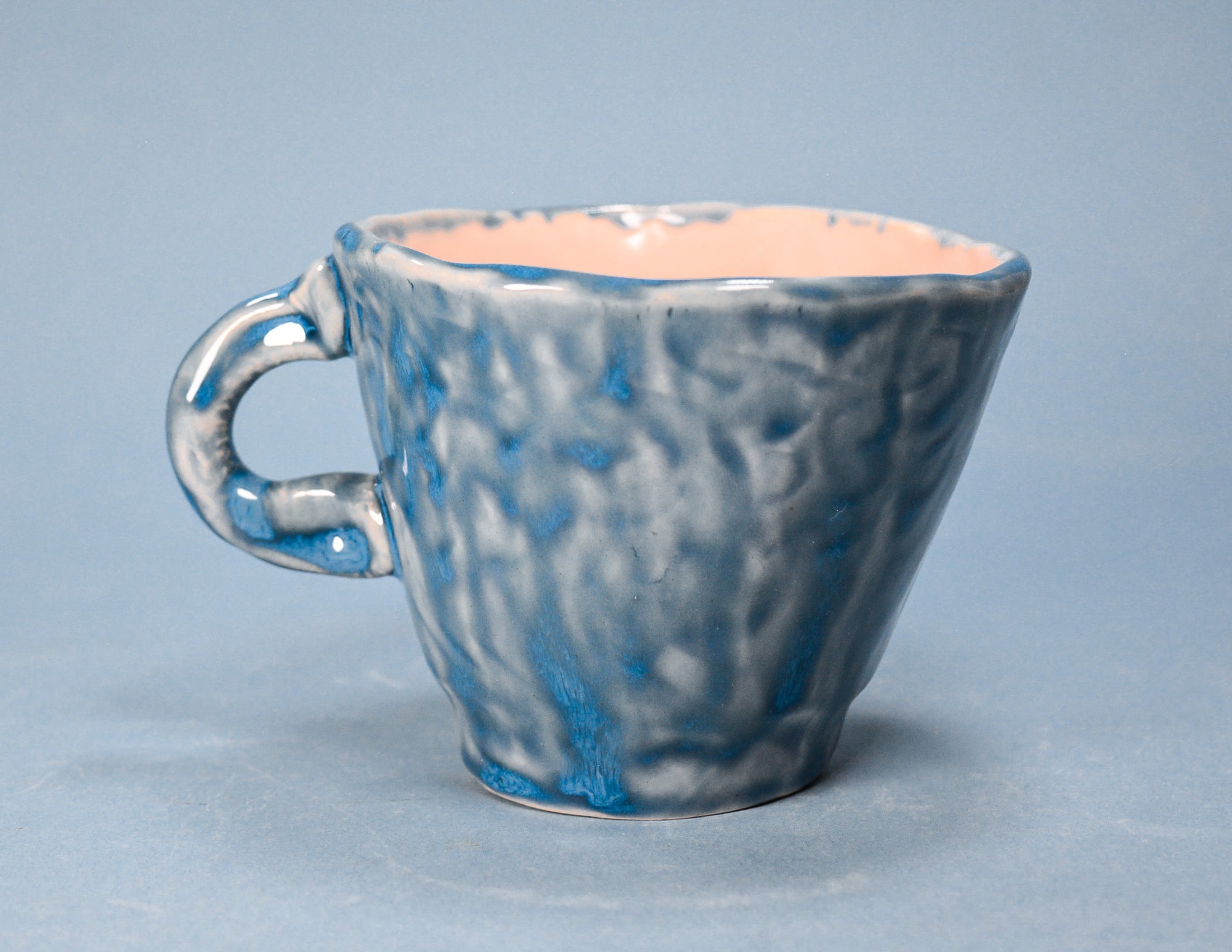 A handmade ceramic coffee cup with a textured, mottled blue and white glaze, featuring a light peach-pink interior, set against a solid light blue-gray background.