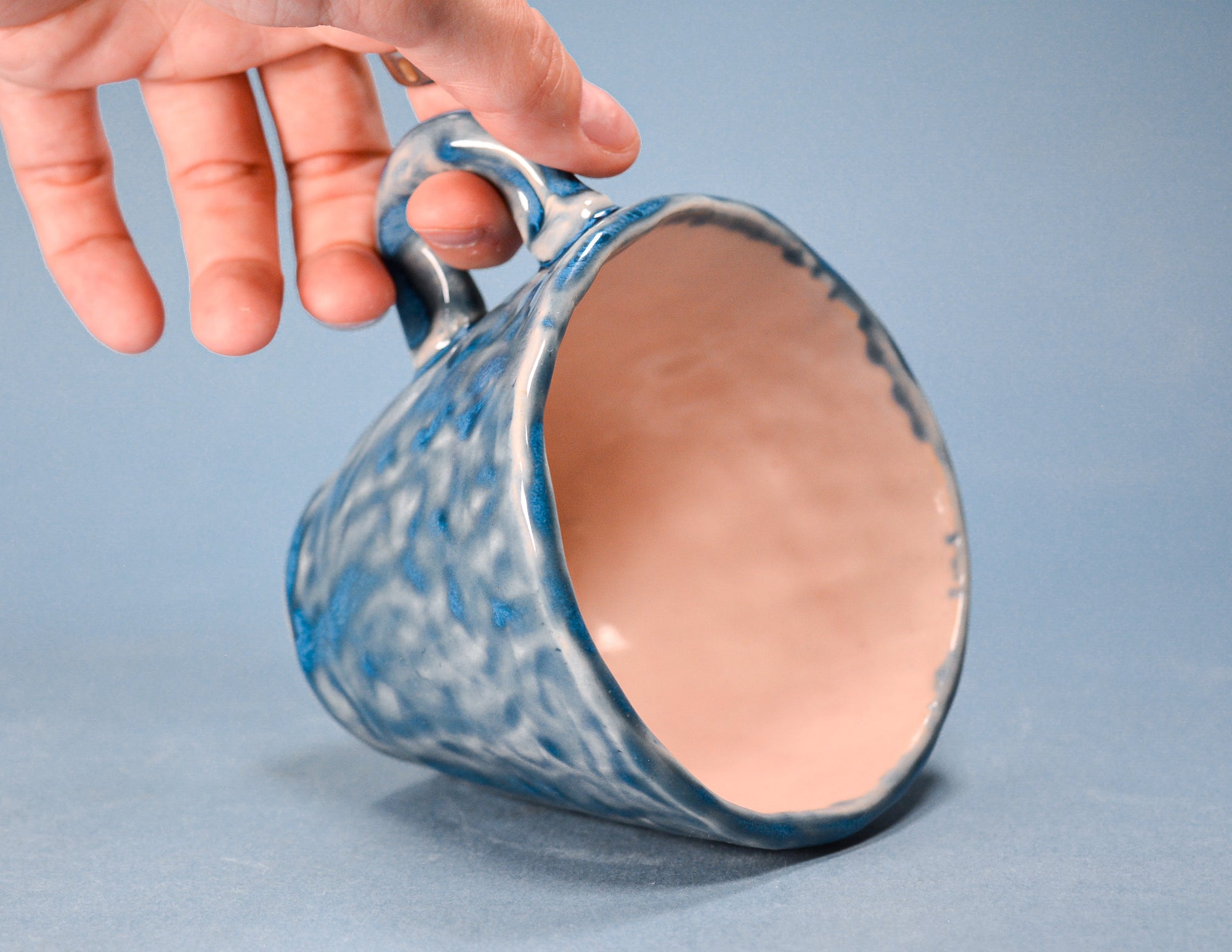 A ceramic mug with a blue and white marbled glaze on the exterior and a warm pinkish-beige interior, held by a hand against a solid blue-gray background.