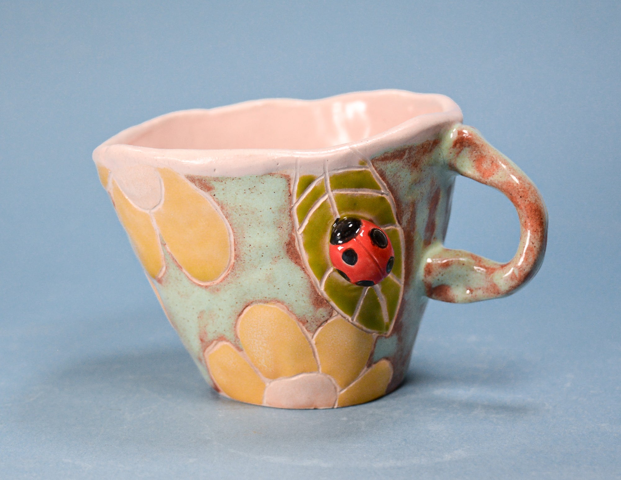 A handmade ceramic teacup with a whimsical design featuring a green leaf with a red ladybug, yellow flower petals, and a pale pink interior, set against a solid blue background.
