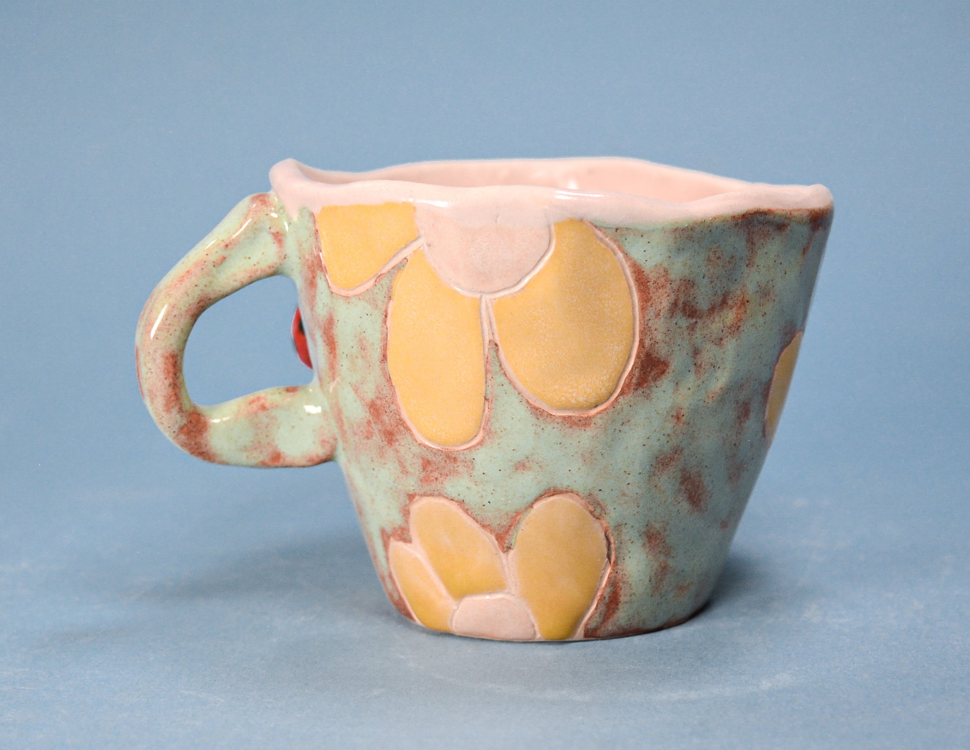 A handmade ceramic cup with a light blue-green glaze, featuring raised yellow and pale pink flower motifs and a speckled brown texture, set against a solid blue background.