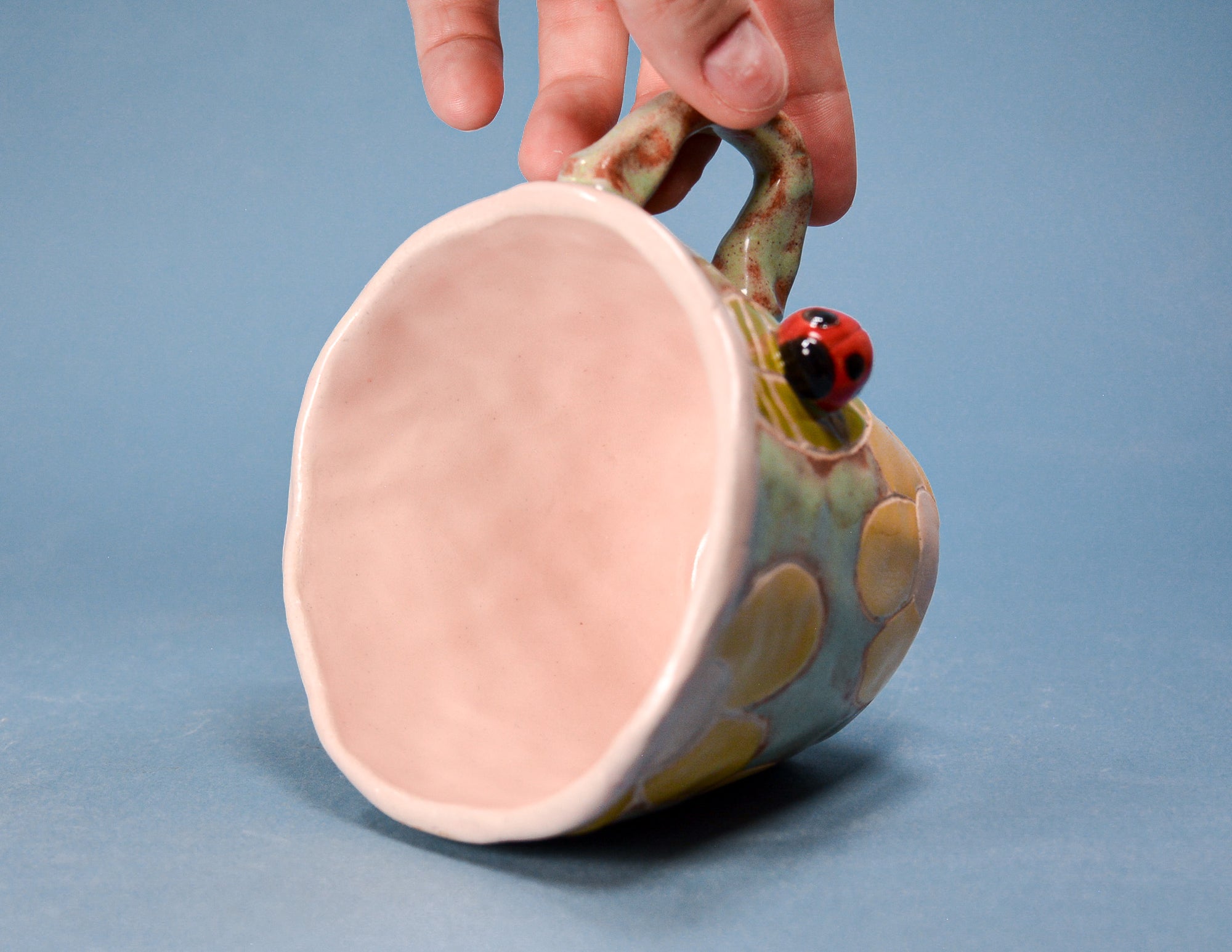 A hand holds a handmade ceramic mug with a light pink interior and a green, yellow, and blue patterned exterior featuring a small ladybug decoration on the handle.