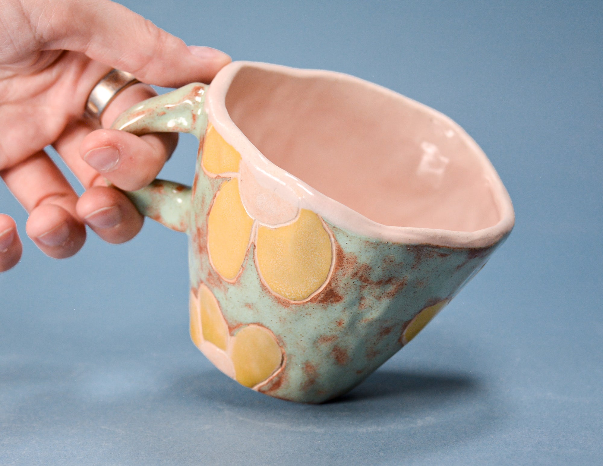 A handmade ceramic mug with a light blue-green glaze, yellow circular patterns, and a pink interior, held by a hand on a blue background.