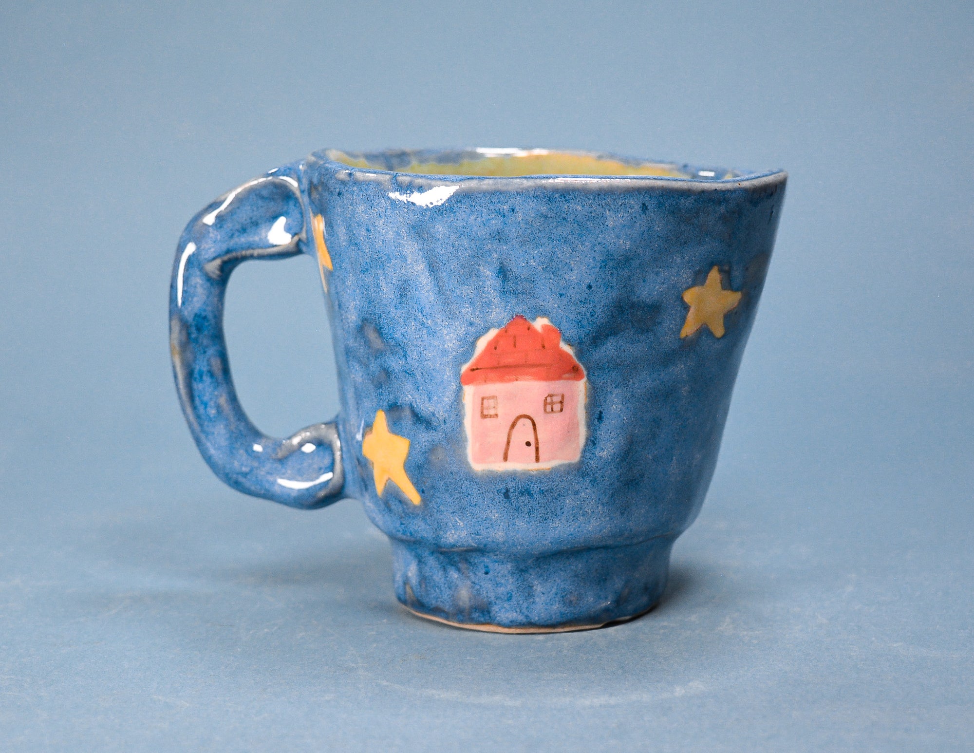 A handmade ceramic mug with a textured blue glaze, decorated with gold stars and a small pink house with a red roof, set against a solid blue background.